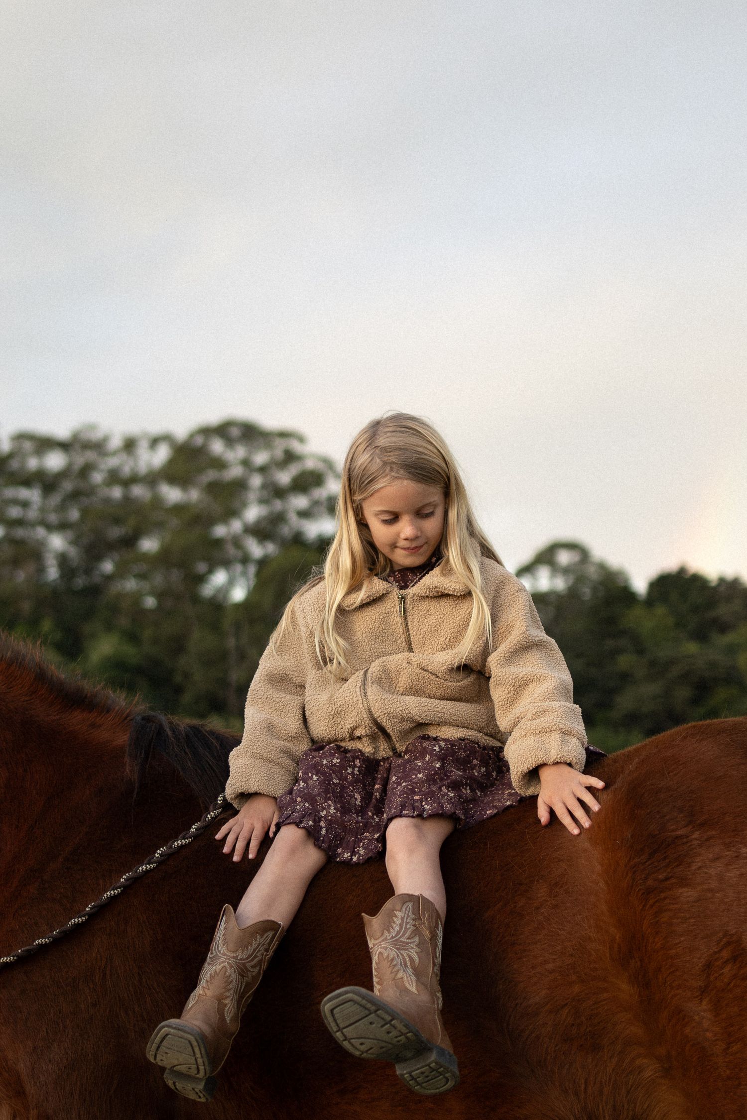 Young girl with long blonde hair in a fluffy beige jacket and cowboy boots sits on a brown horse against a soft evening sky.