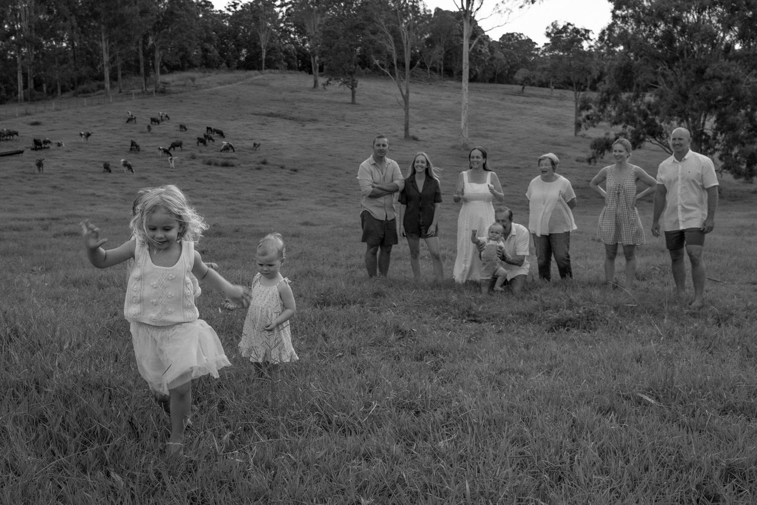 Two young girls run laughing across a grassy hillside while a family of adults watches in the background in this black and white photo.