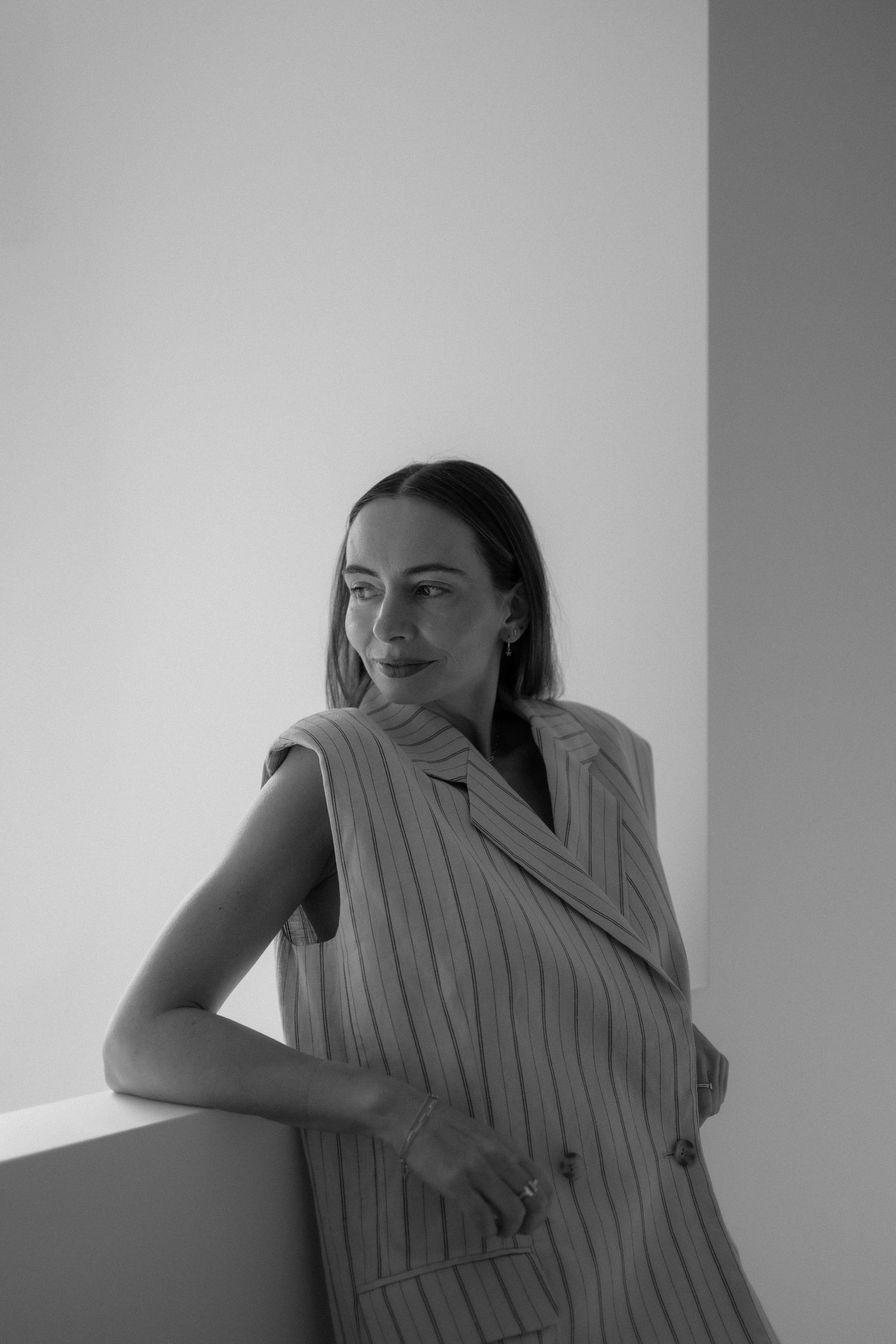 Black and white portrait of a smiling woman in a pinstriped sleeveless blazer leaning on a ledge and looking away.