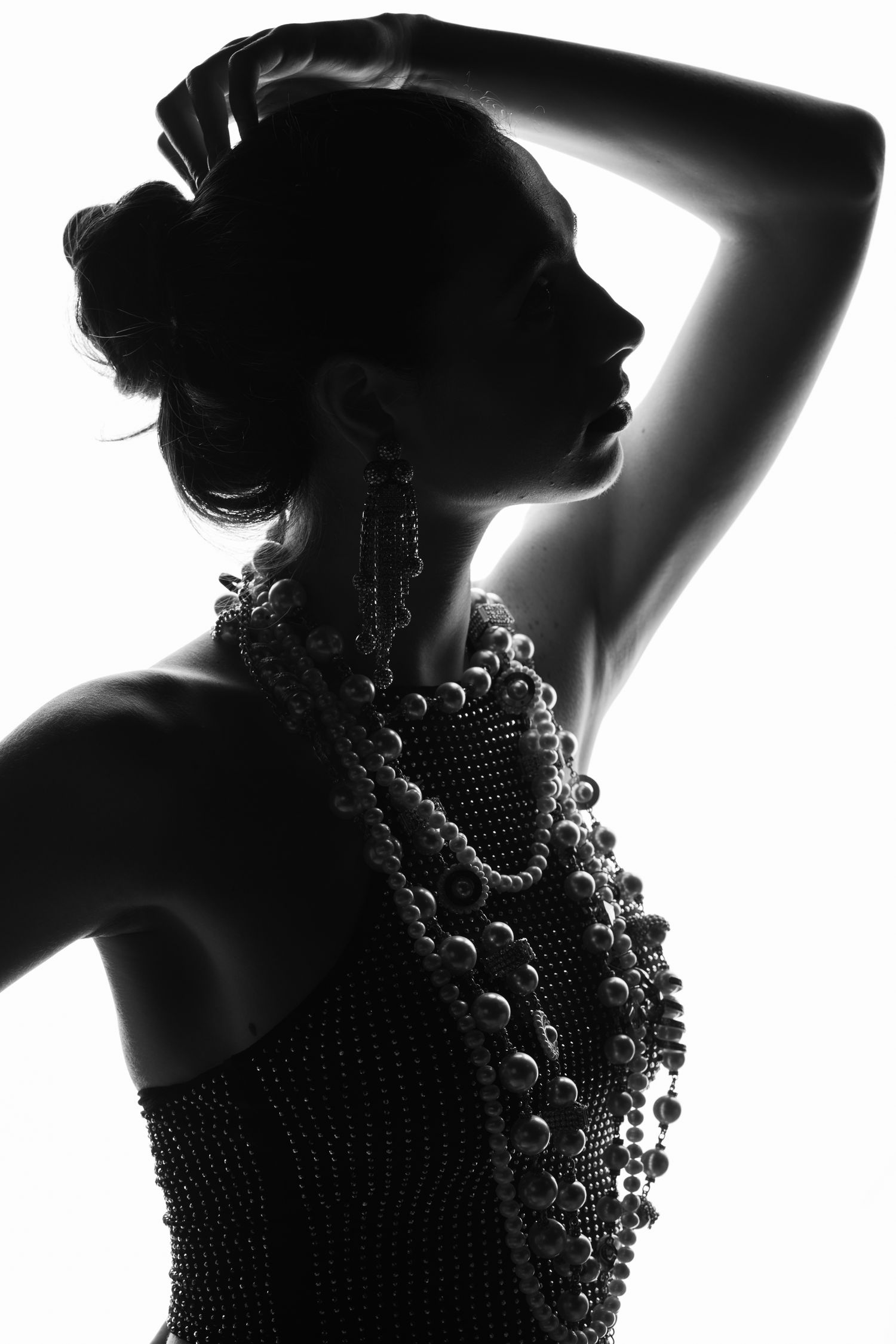 Elegant woman in dramatic black and white silhouette portrait, side profile with raised arm, wearing statement beaded jewelry and bun hairst