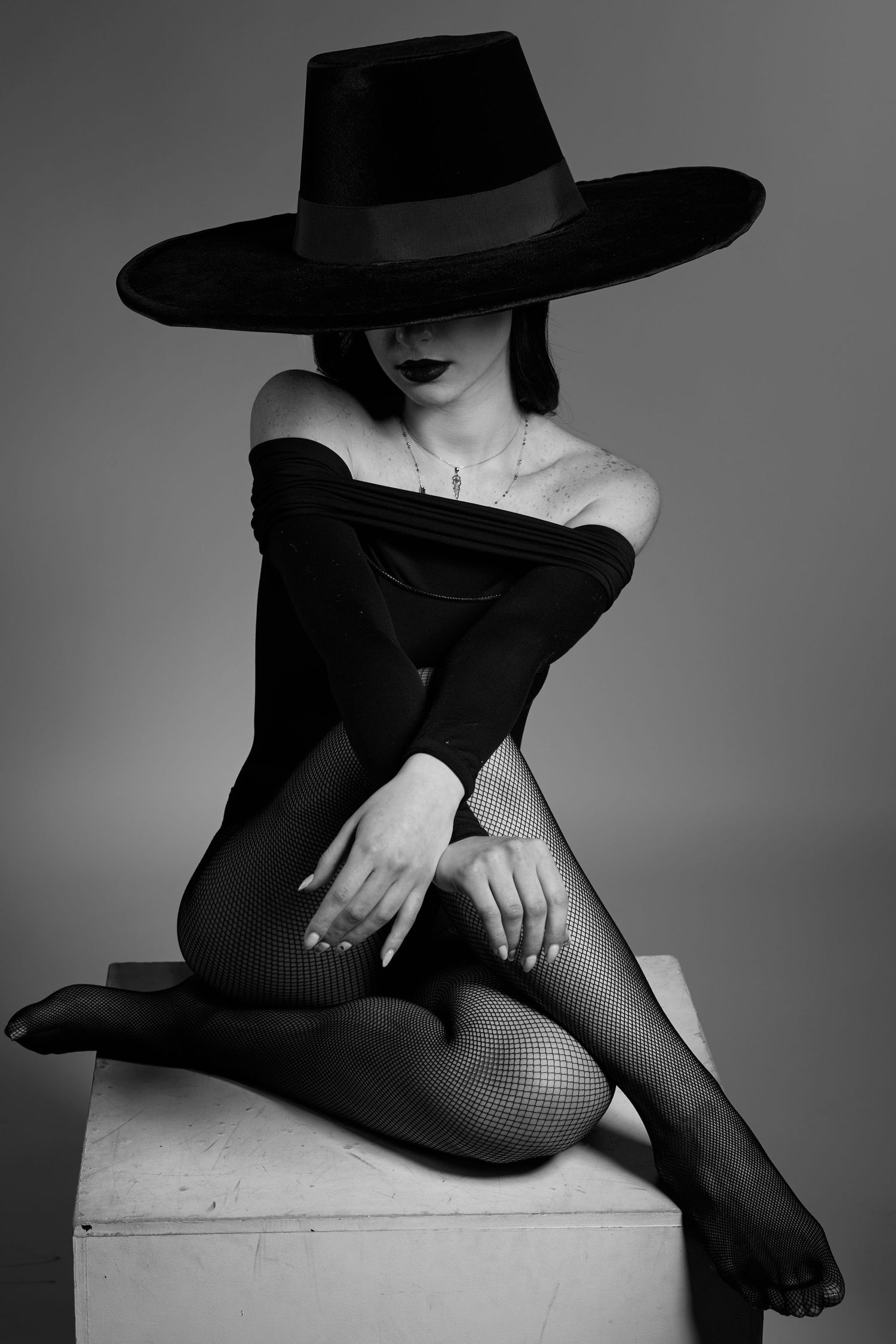 Black and white fashion portrait of seated model in wide-brim hat, minimalist and elegant composition.