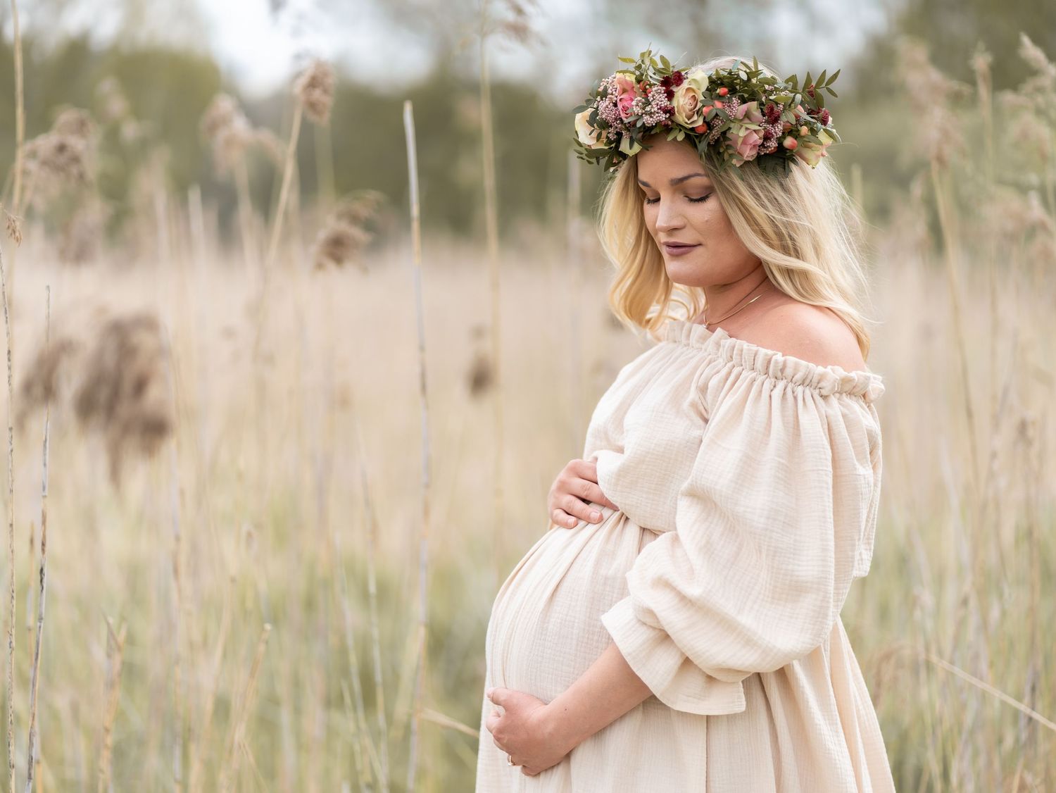 Gravid kvinna med blomsterkrans i beige bohemisk klänning fotograferad utomhus i somrigt fält. Gravidfotografering i Stockholm.