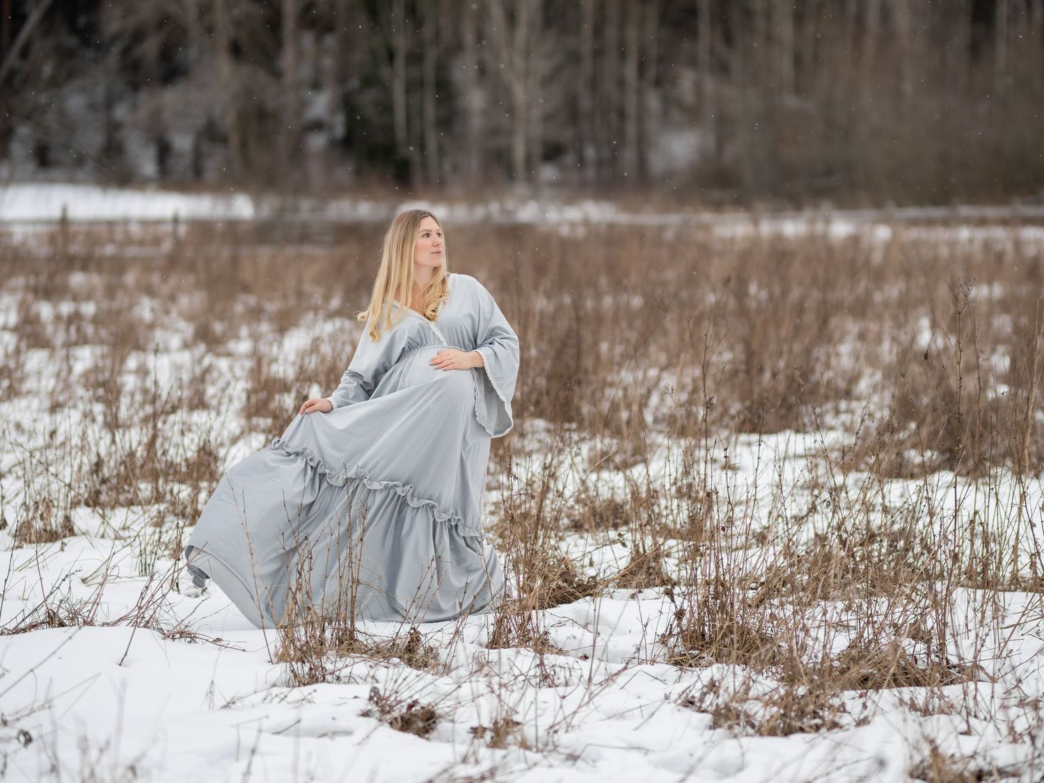 Gravid kvinna i ljusblå klänning med spetsdetaljer fotograferad i vintrigt landskap med snö och torrt gräs, gravidfotografering i Stockholm.