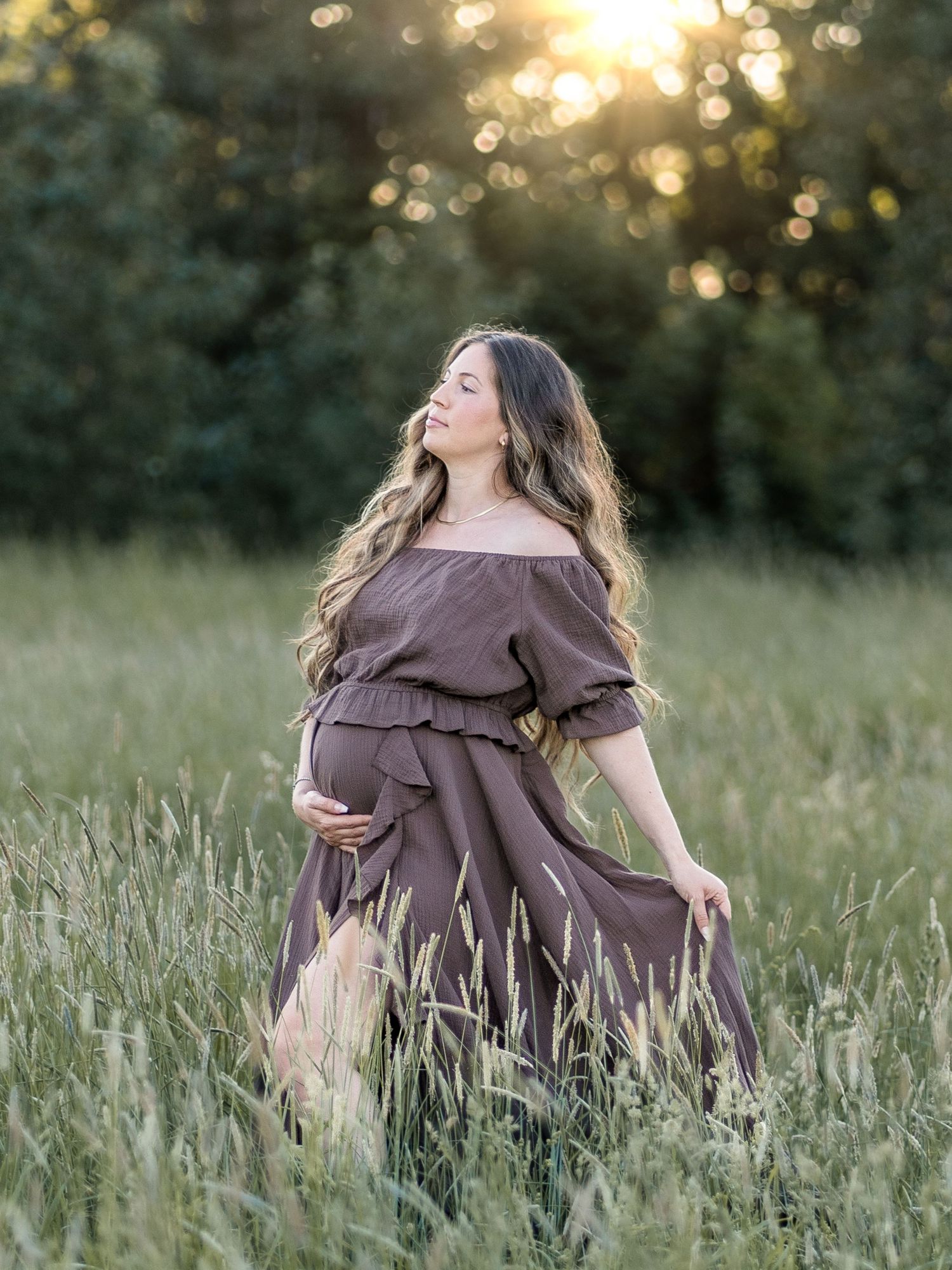 Gravid kvinna i brun bohemisk klänning fotograferad i grönskande fält i kvällsljus. Gravidfotografering utomhus i Stockholm.