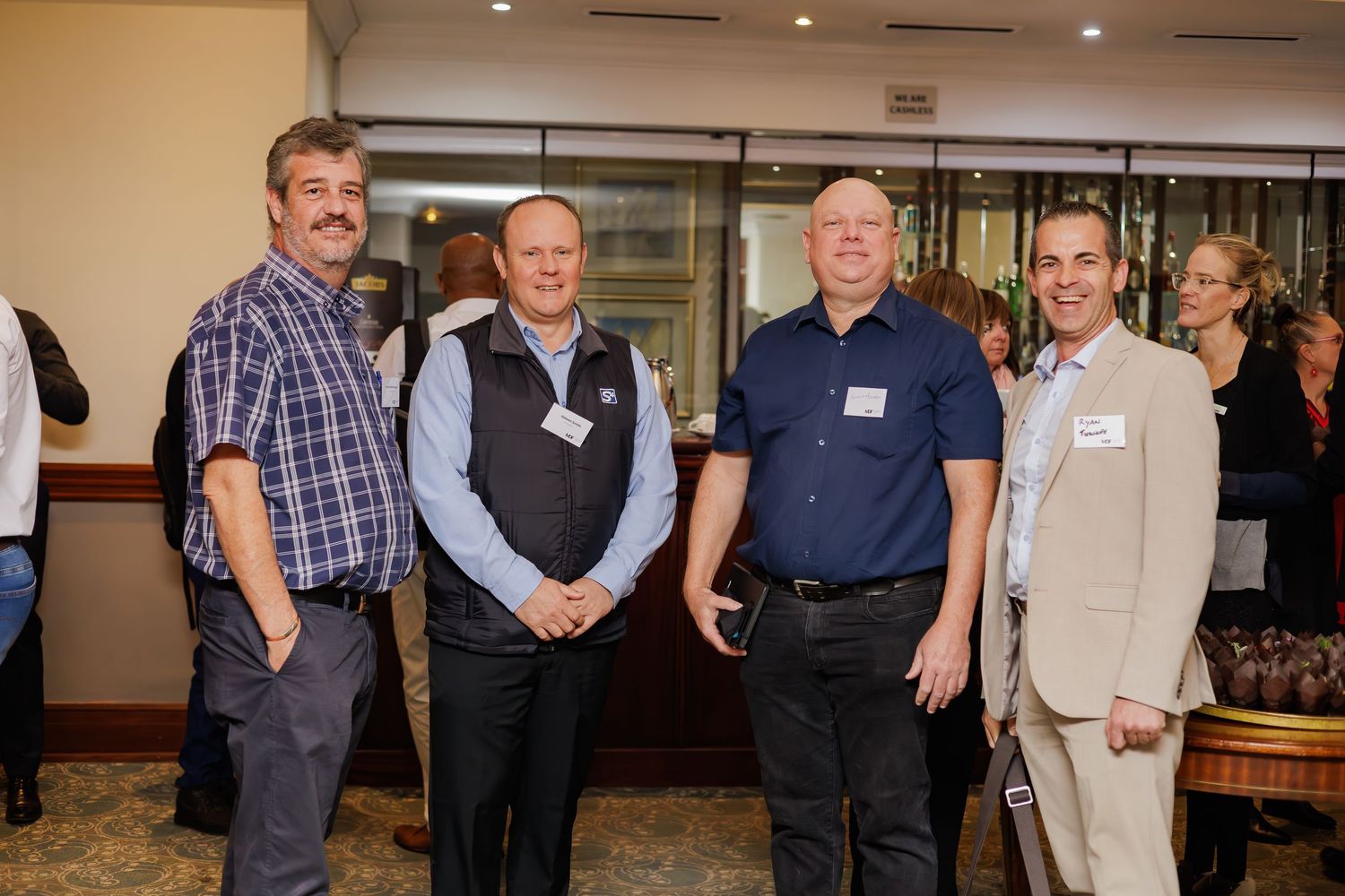 Four men wearing name tags smiling together at a corporate networking event in a hotel venue.