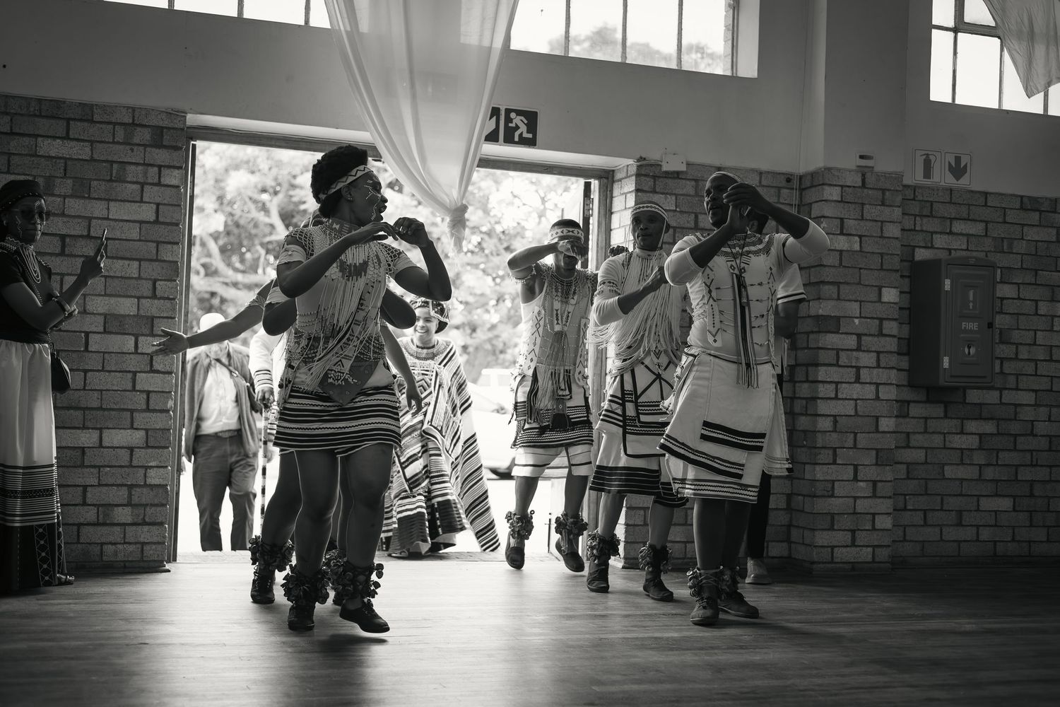 A black and white photo of people performing traditional African dance in a brick building.