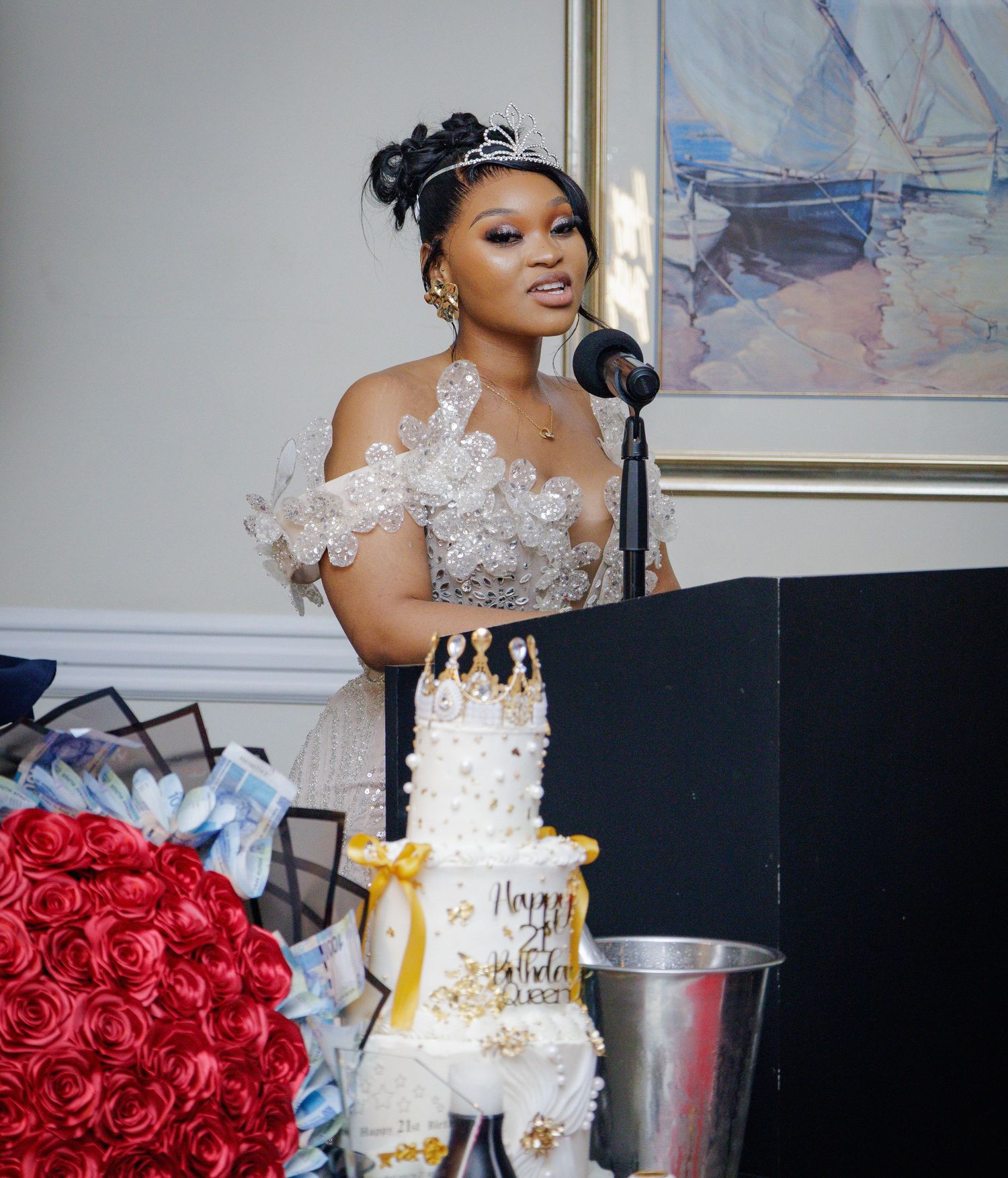 Elegant woman in white gown with tiara speaks at podium beside a tiered Happy Birthday cake.