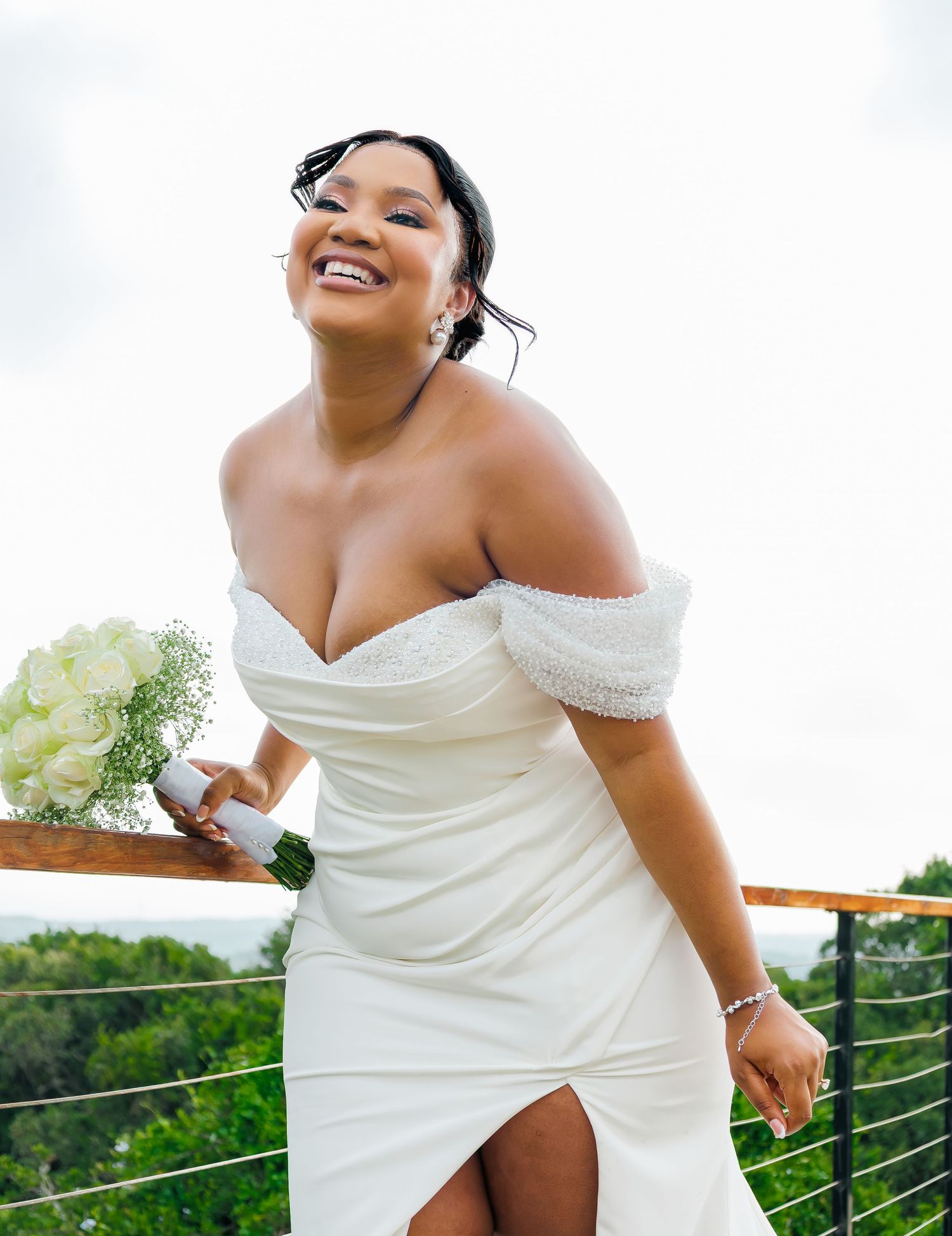 A radiant bride in an off-shoulder white wedding dress holds a bouquet while smiling on a balcony.