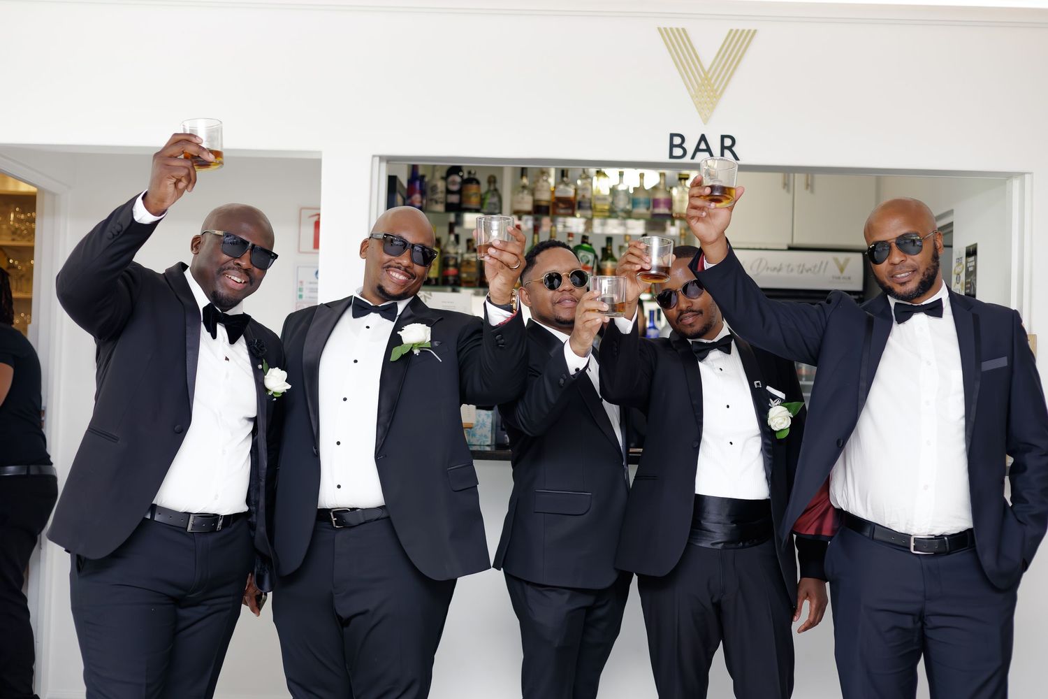 Groom & groomsmen in black tuxedos raising glasses in a toast at a wedding bar celebration.