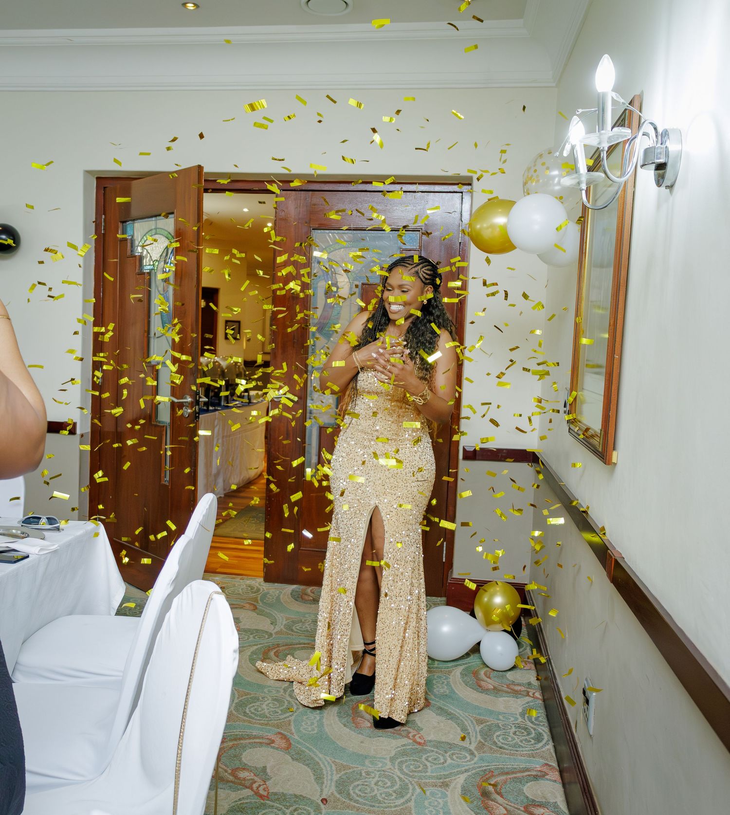 Elegant woman in gold sequin slit gown standing amid golden confetti shower at a celebration event.
