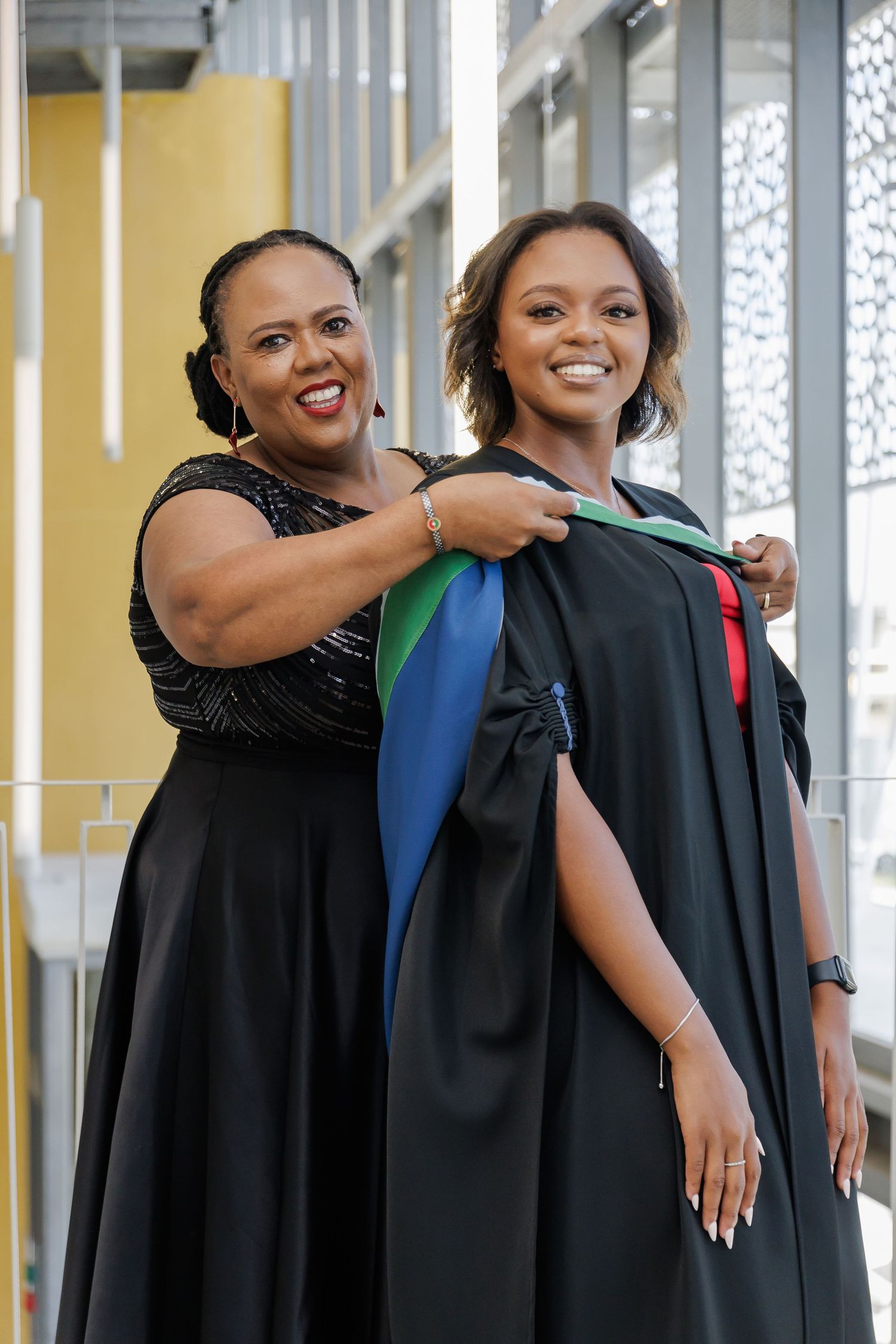A proud Black mother adjusts her daughters graduation hood as they smile together inside a modern building.