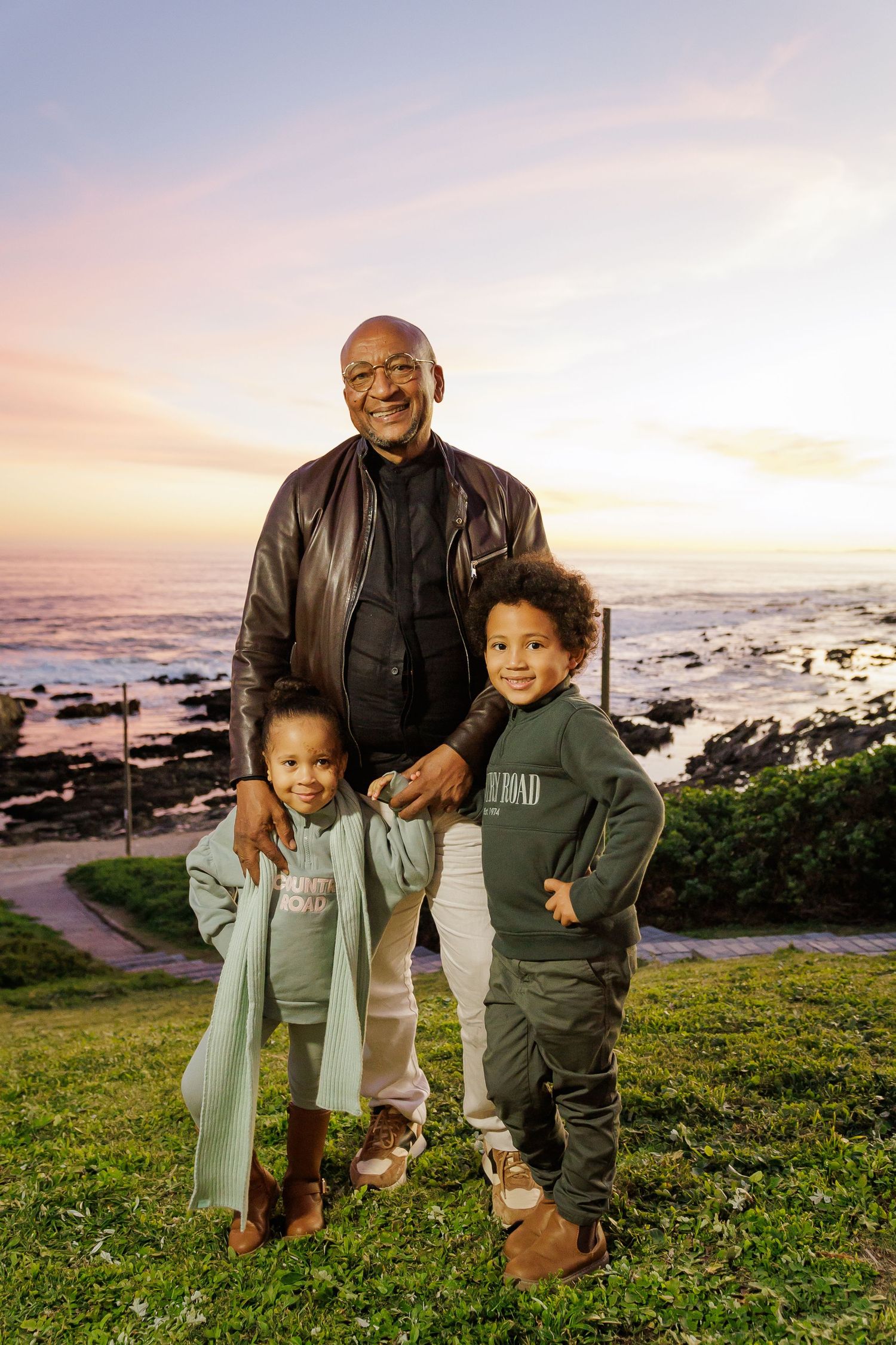 Proud grandfather standing with two young grandchildren on coastal lawn during a colorful sunset.