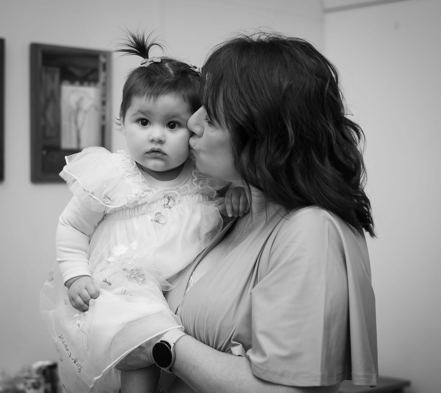 Woman tenderly kissing baby girl in white dress in black and white portrait.