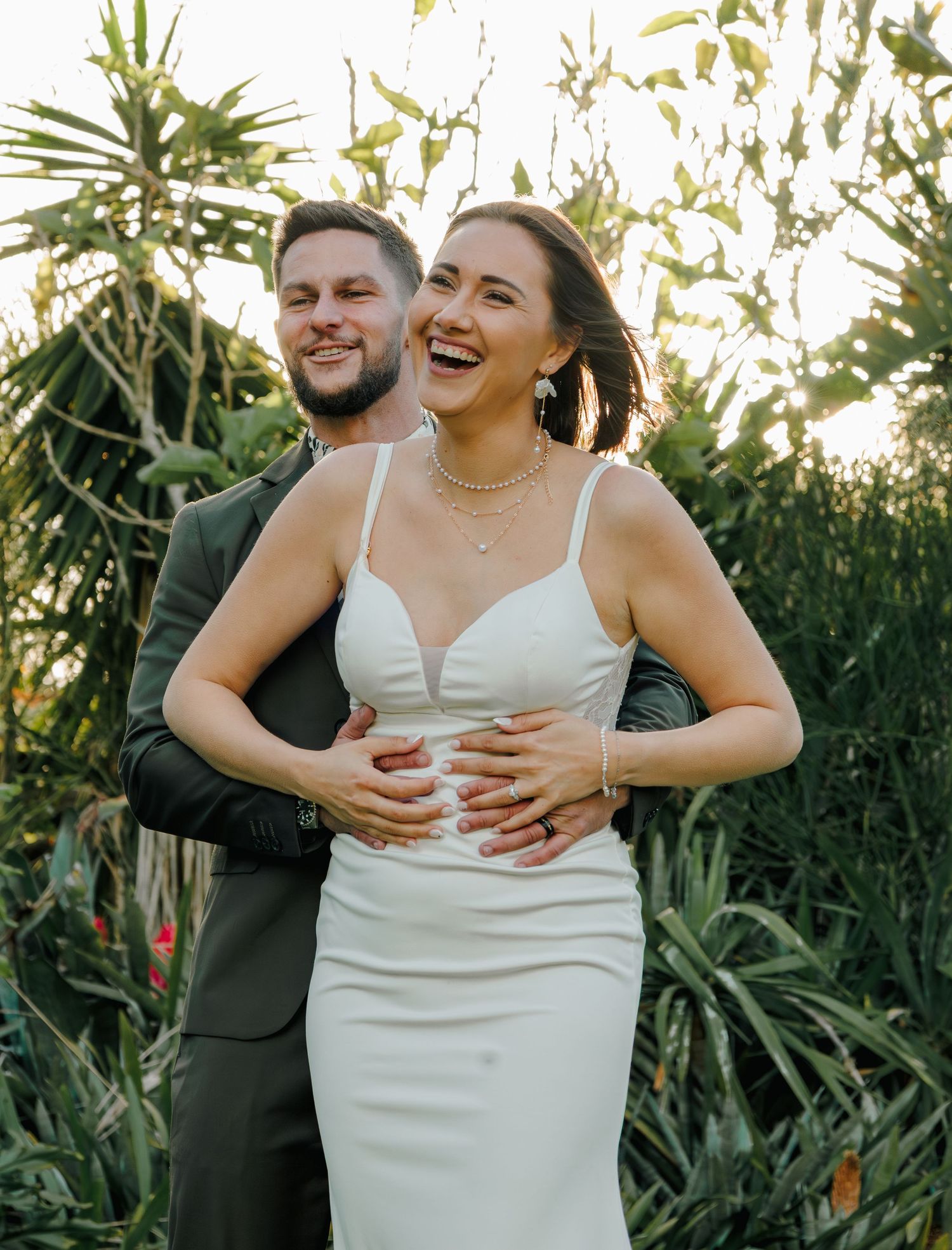 Laughing bride in white fitted gown embraced from behind by smiling groom in dark suit outdoors.