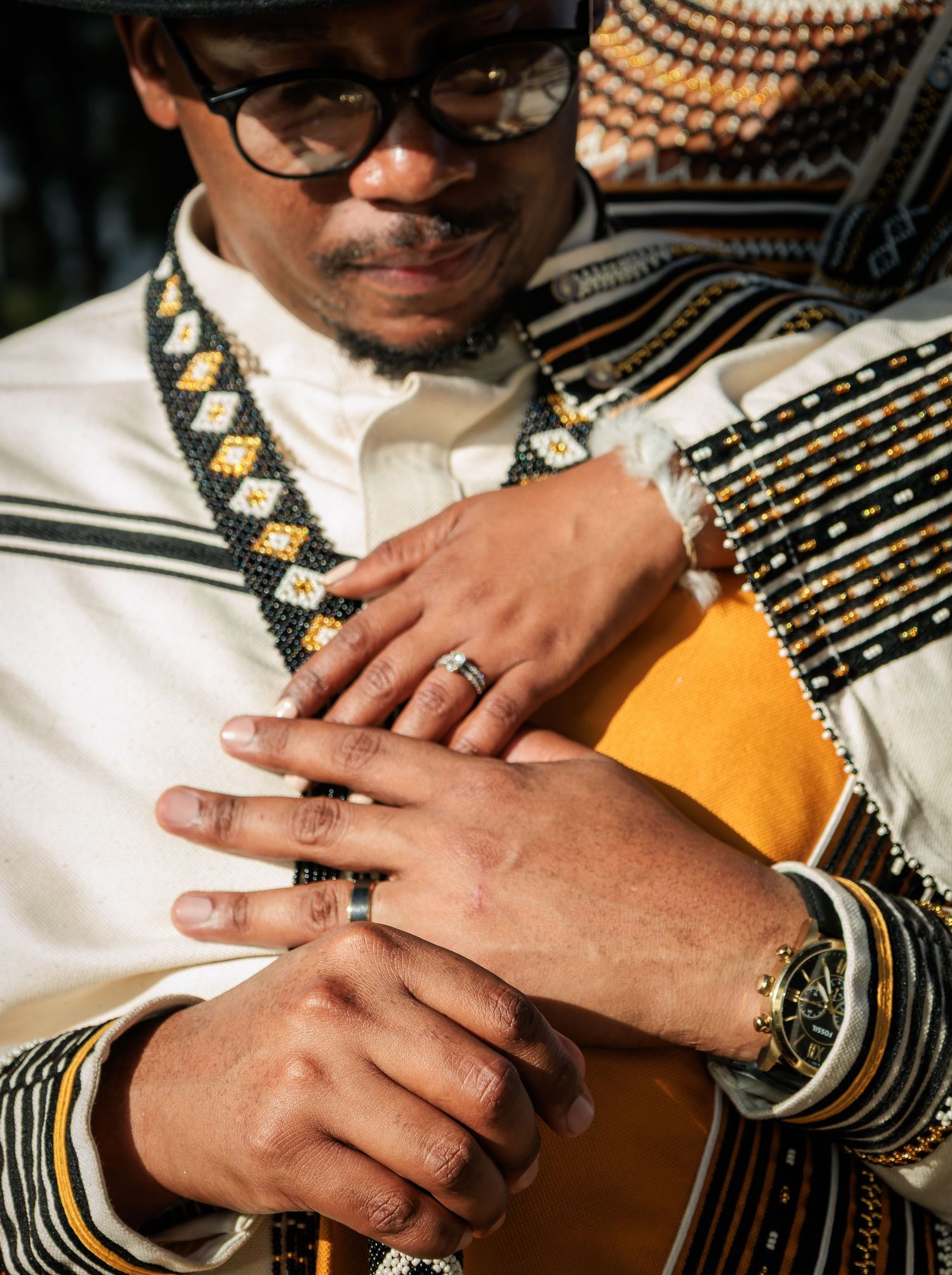 Close-up of couple's hands intertwined, wearing wedding rings and traditional Xhosa patterned attire.