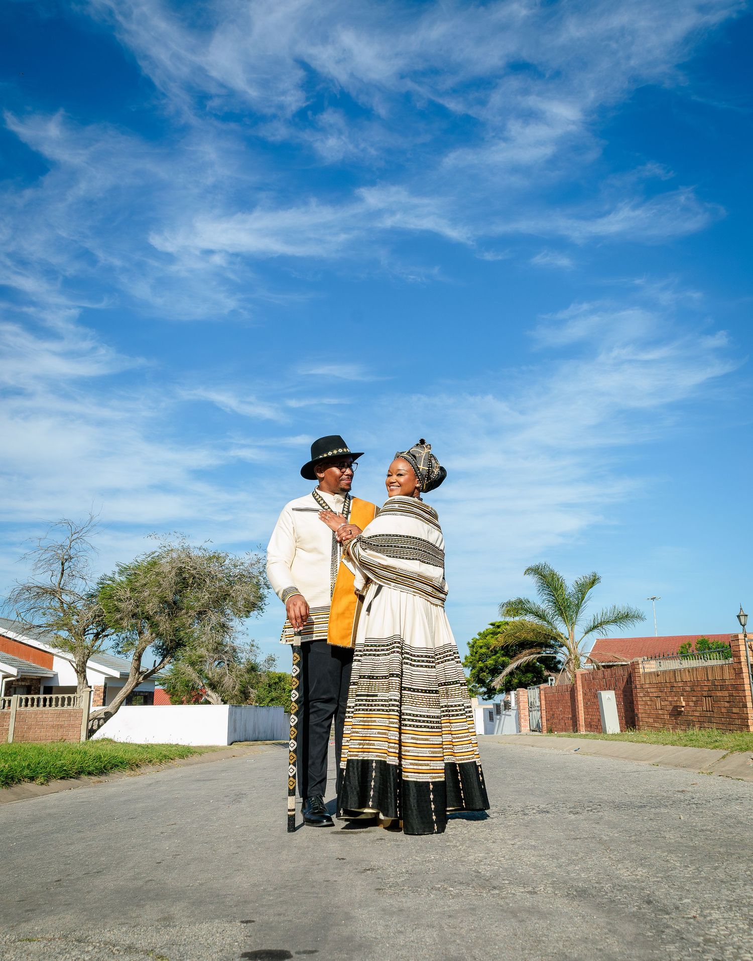 Couple standing on street in traditional Xhosa clothing under blue sky with scattered clouds.