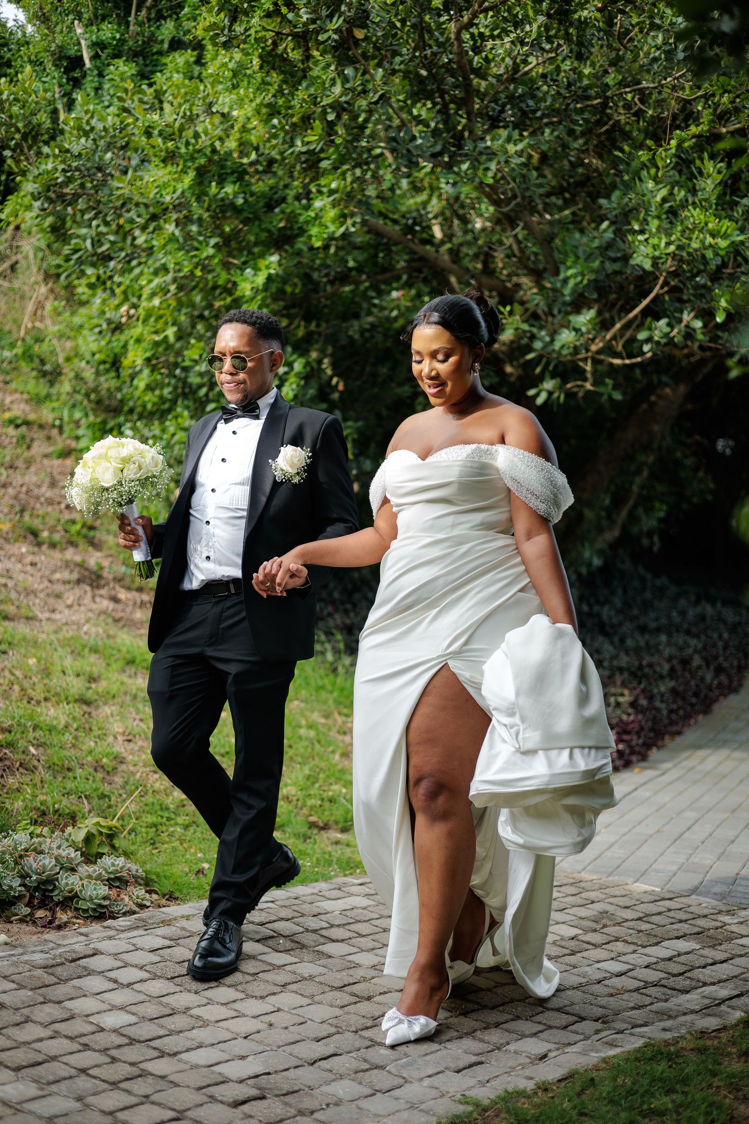 Smiling bride in off-shoulder gown walks with sunglasses-wearing groom holding bouquet outdoors.