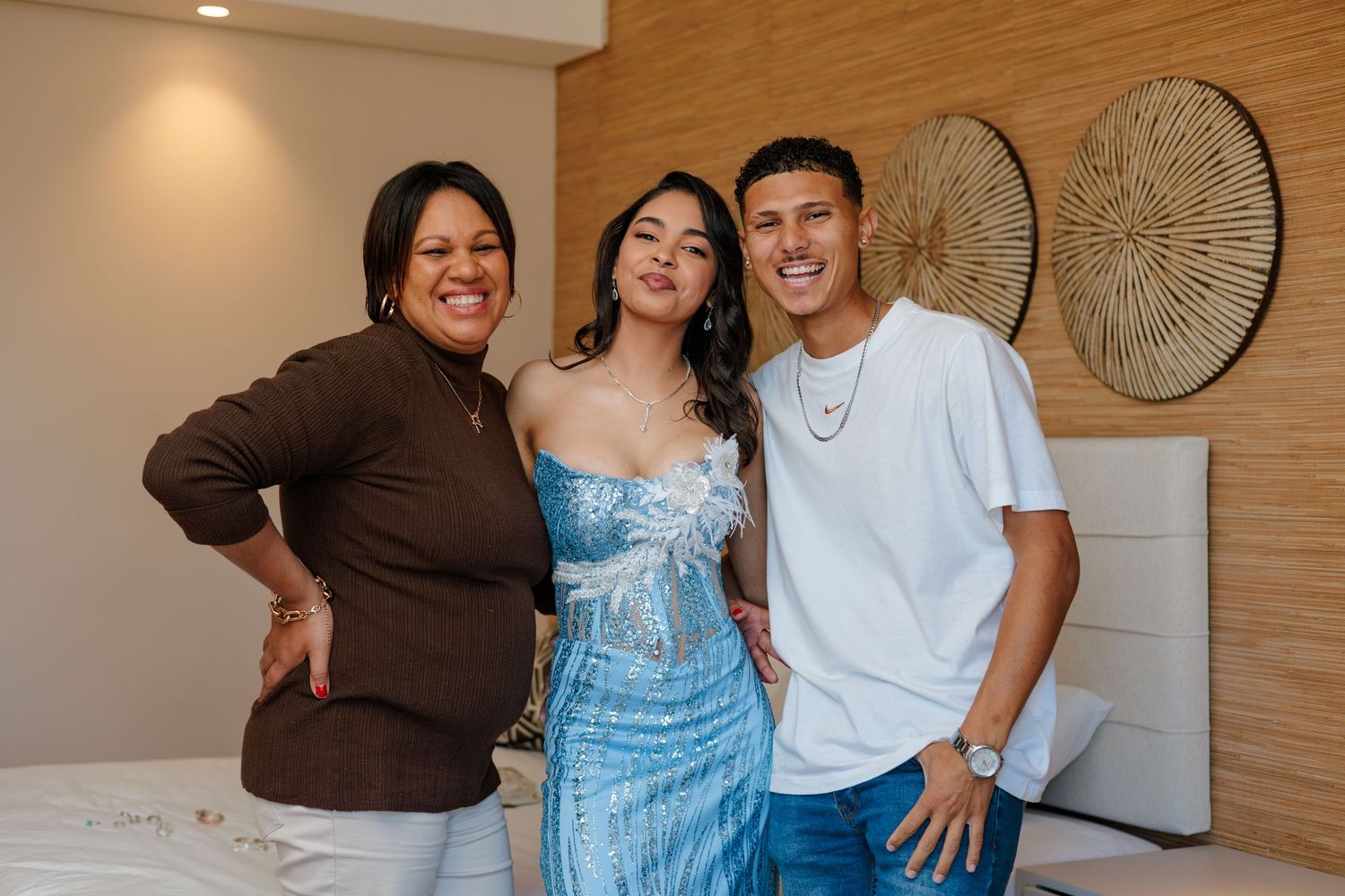 Three people smiling and posing together in a stylish room, woman in blue sequin dress flanked by two others.