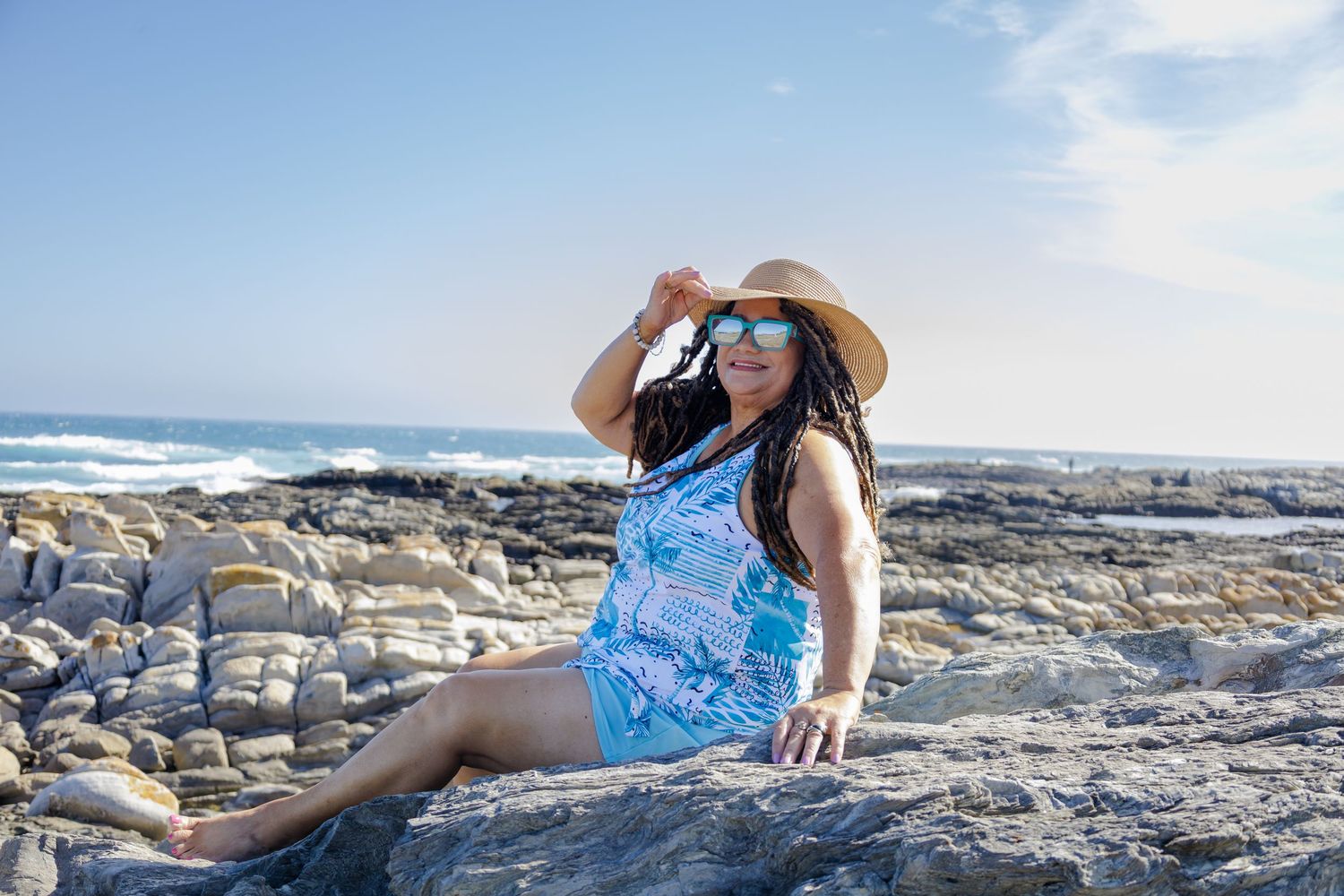 Woman in blue swimsuit, straw hat, and sunglasses posing on rocky beach with ocean waves in background.