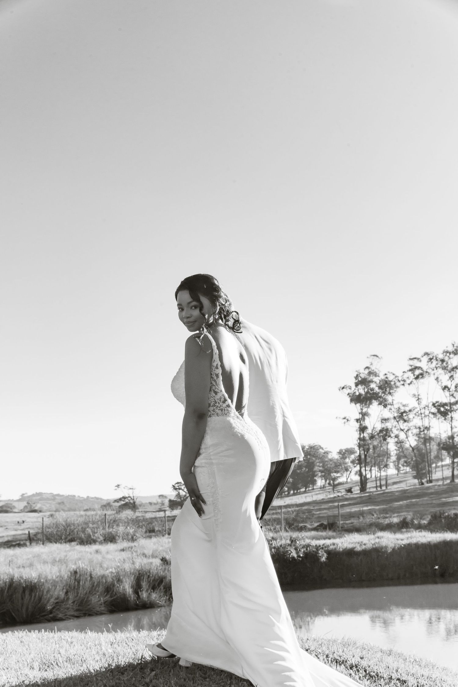 Elegant bride in a fitted white gown smiles over her shoulder during outdoor wedding photos by a pond.