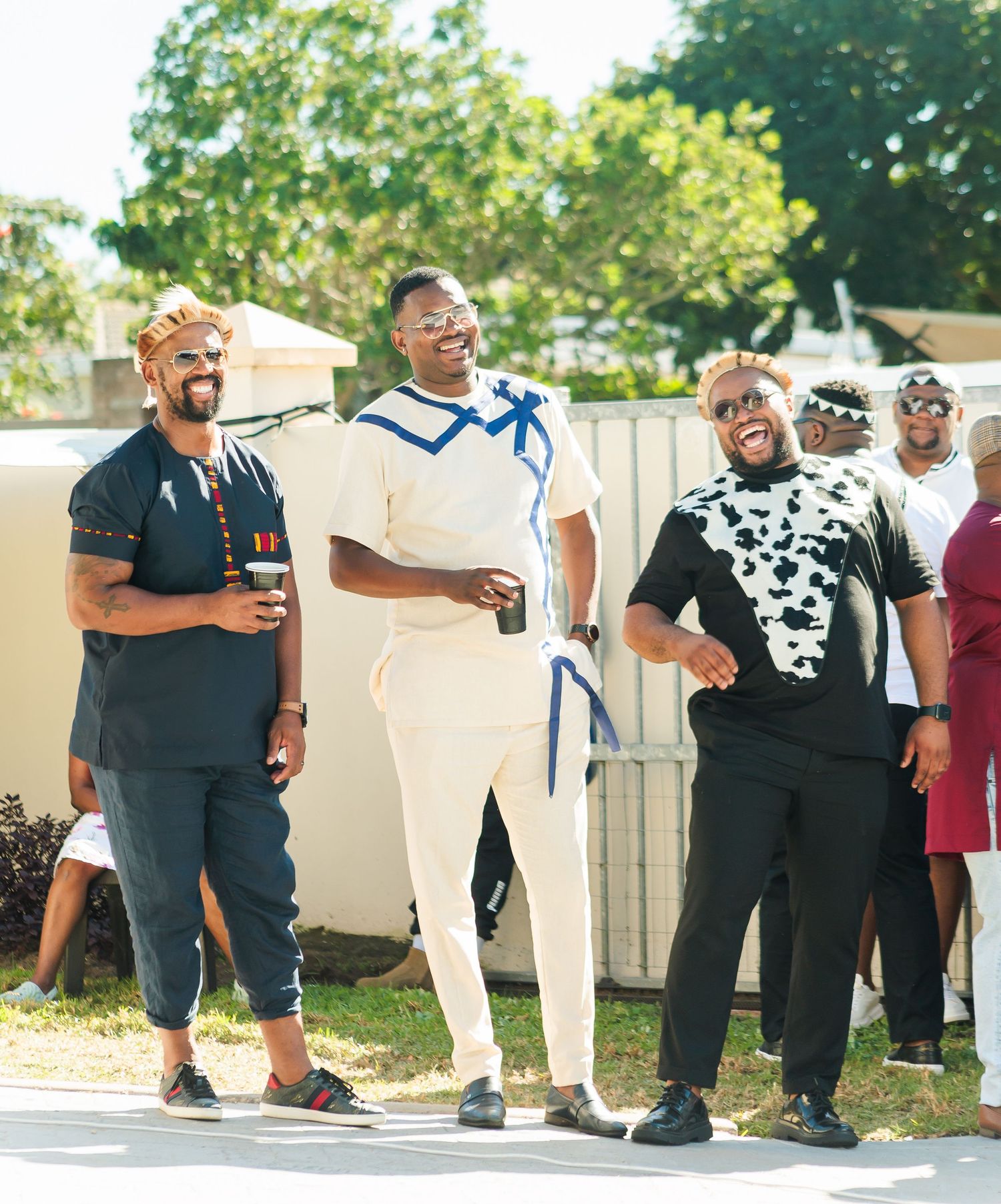 Three stylish men in traditional African outfits laughing and celebrating outdoors at a wedding event.