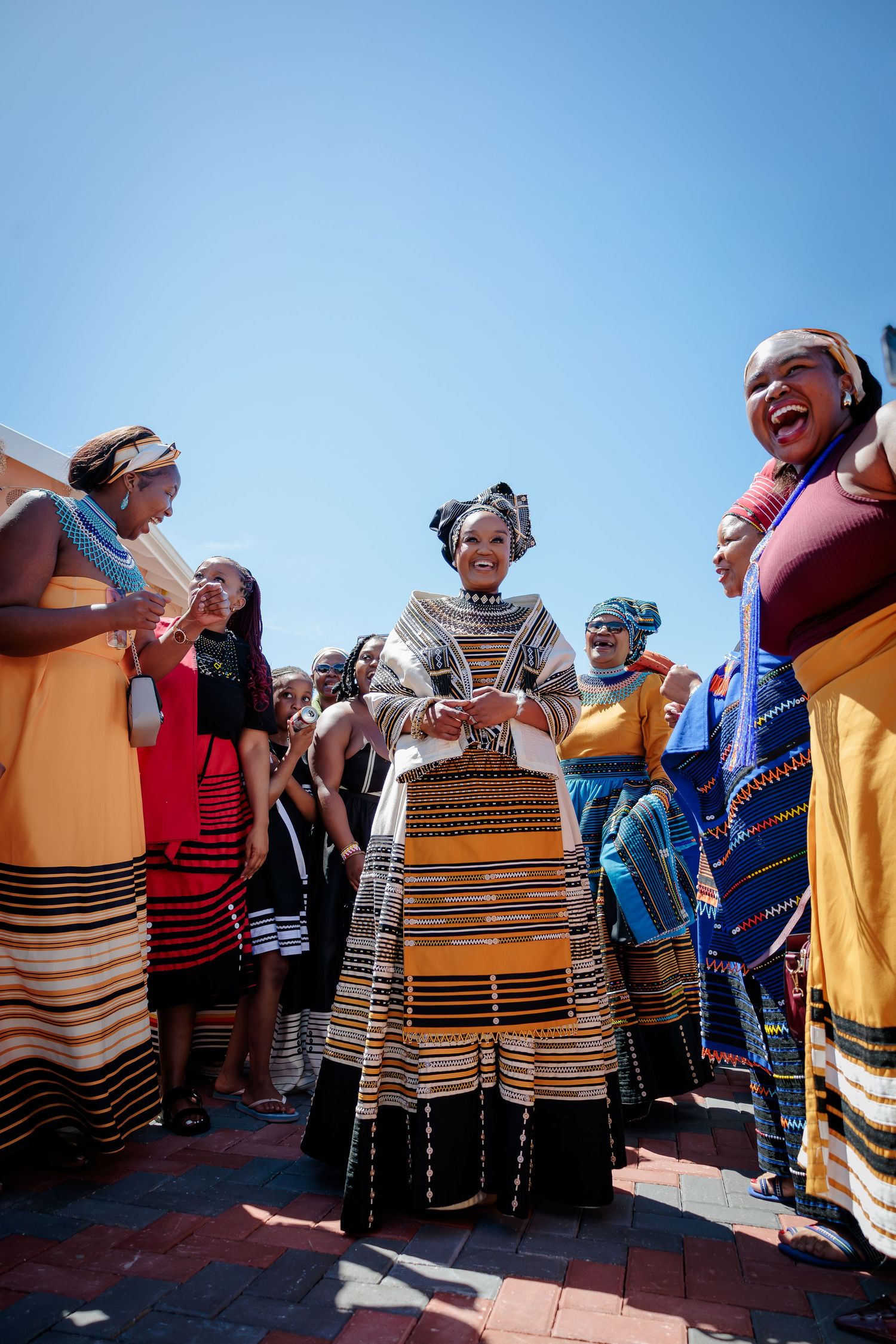 Bride in traditional Xhosa umbhaco dress stands joyfully surrounded by celebrating women outdoors.