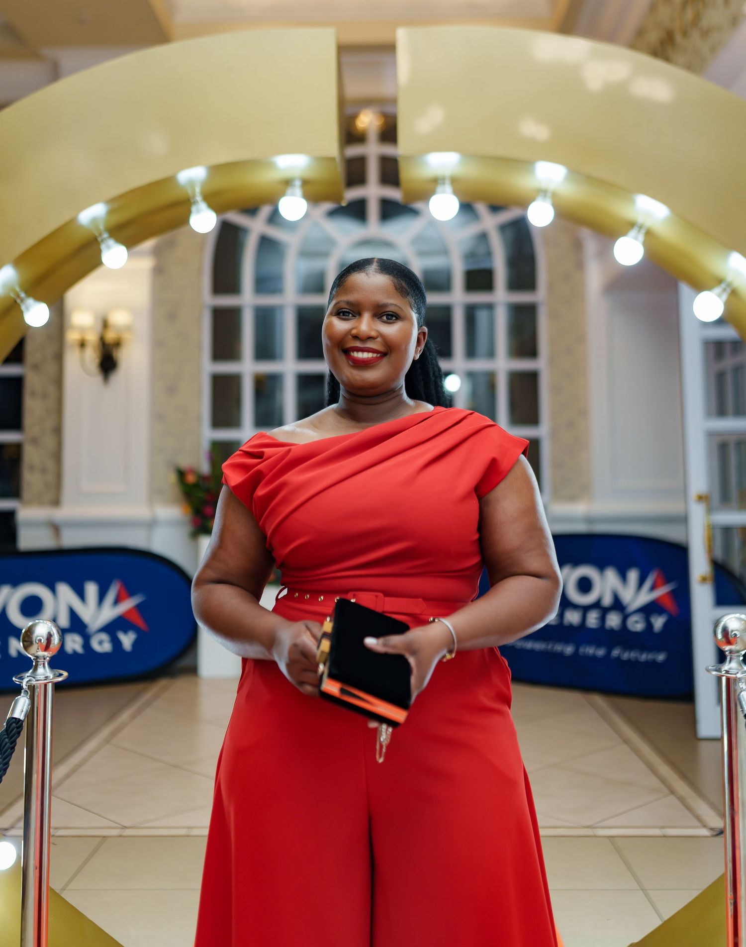 Woman in red off-shoulder jumpsuit posing on orange carpet at Beyond Energy gala.