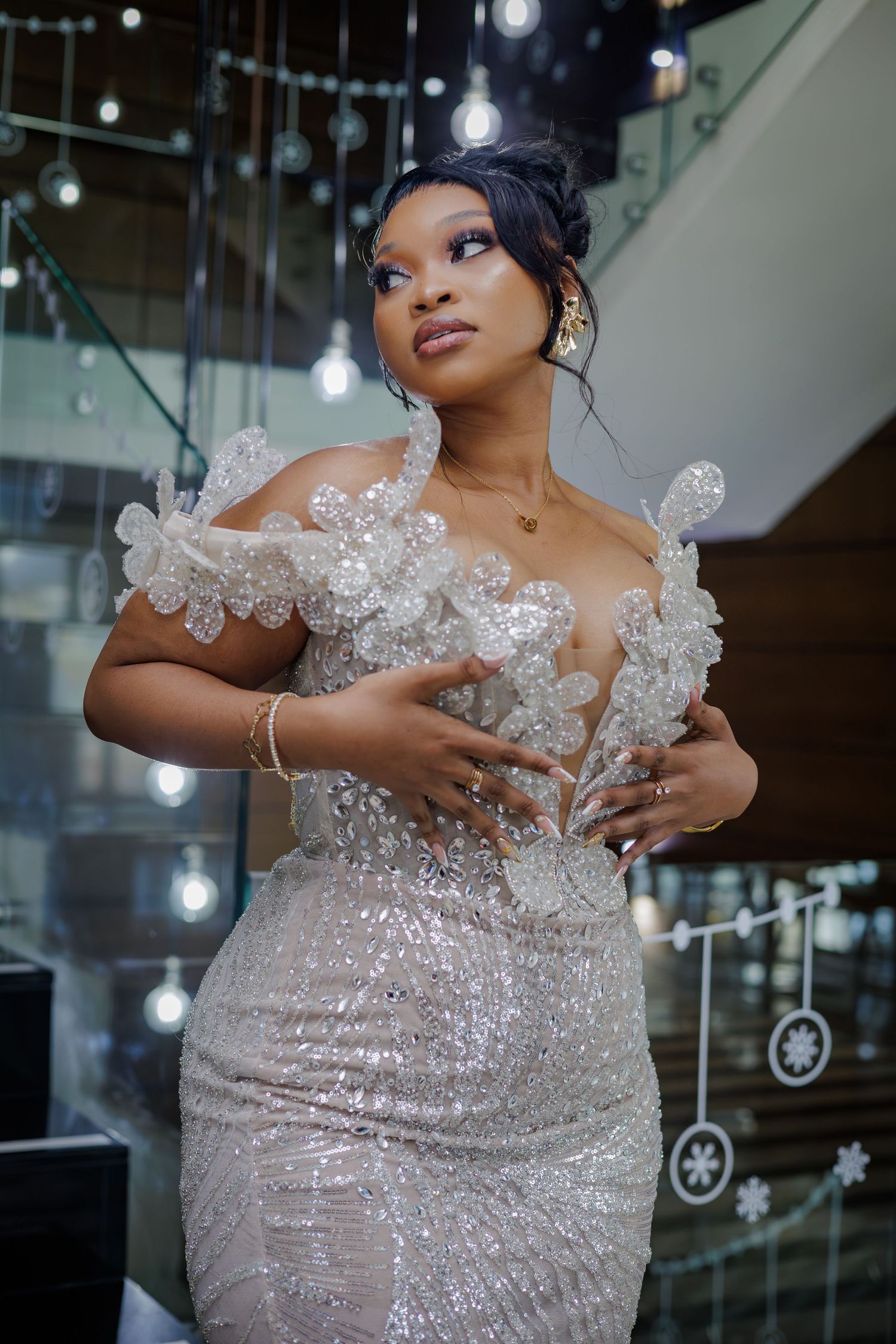 Woman in elegant silver sequined off-shoulder gown with floral details posing near decorative lights.