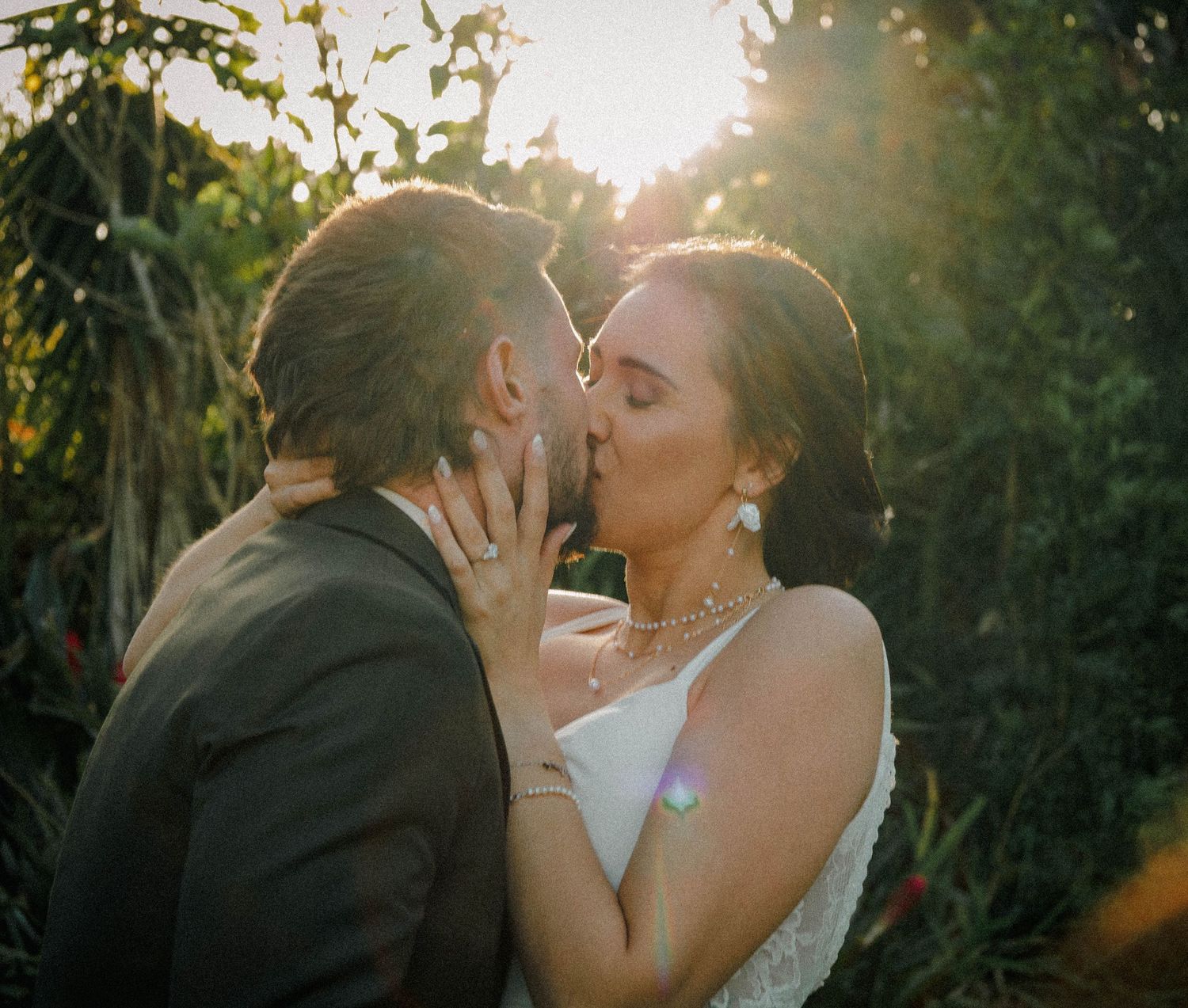 Bride and groom share romantic first kiss bathed in warm golden sunlight among lush garden.