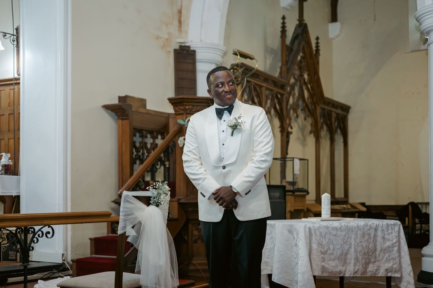 Smiling groom in white tuxedo jacket waits at church altar for bride.