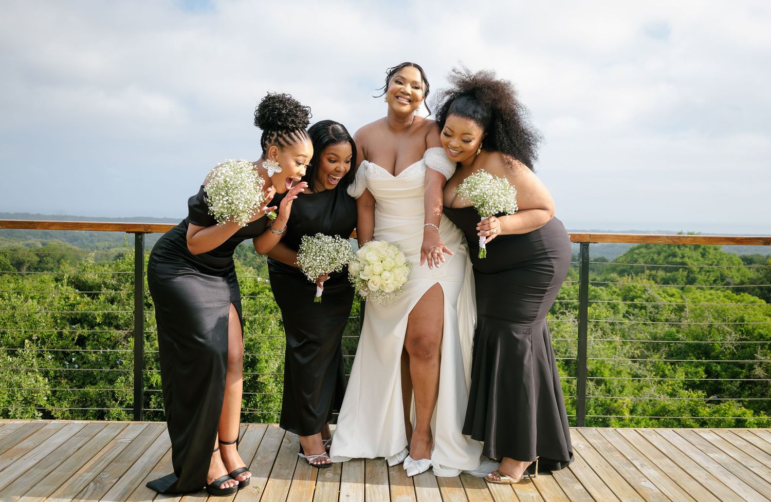 Bride in white gown poses with three bridesmaids in black dresses holding white bouquets outdoors.