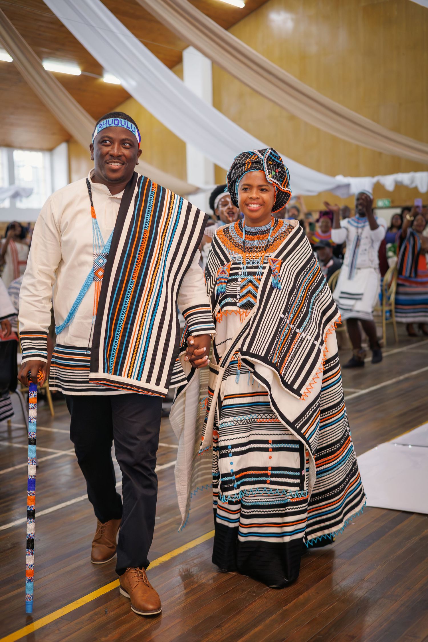 Couple in traditional Xhosa attire walking hand in hand at a cultural wedding ceremony.
