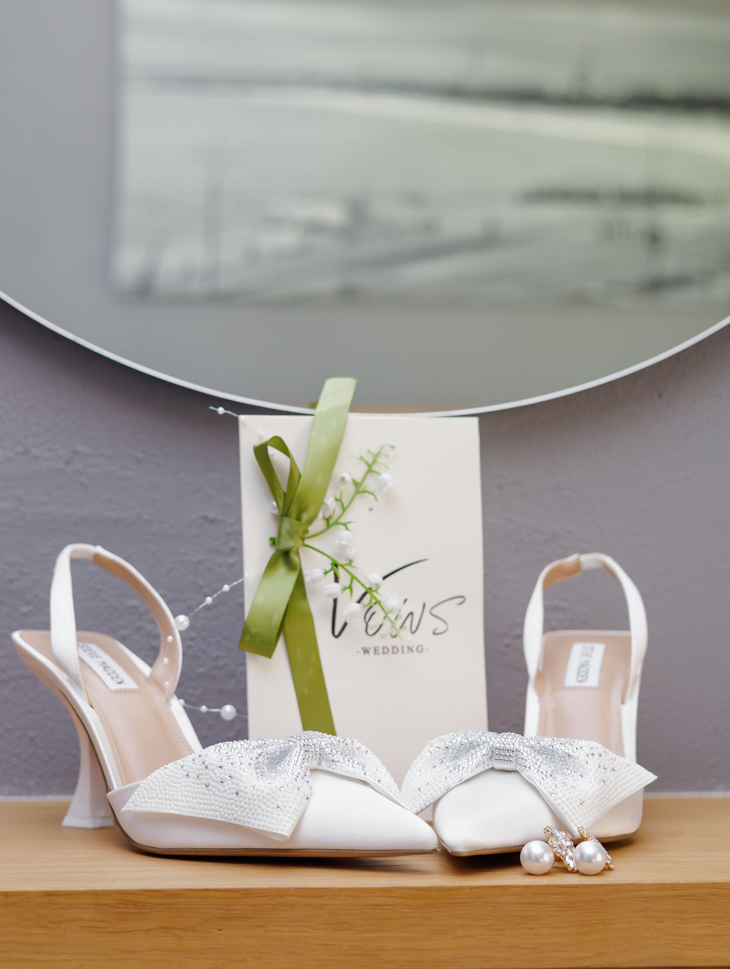 White bridal heels with crystal bow accents displayed beside pearl earrings and a Vows Wedding card.