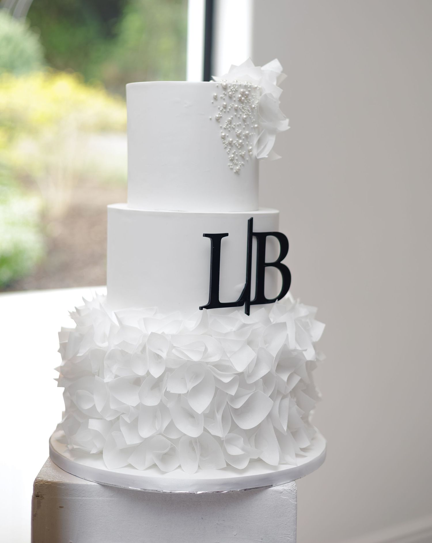 Three-tier white wedding cake with ruffled bottom tier and black monogram letters L and B.