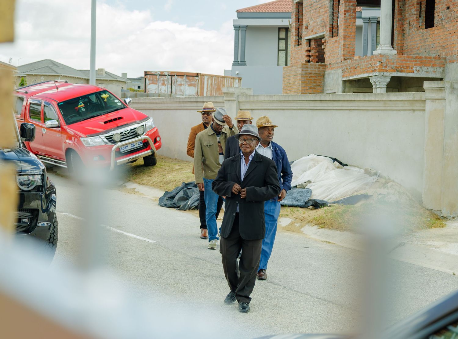 Men in formal attire walking along a street past parked vehicles including a red Toyota Hilux bakkie.