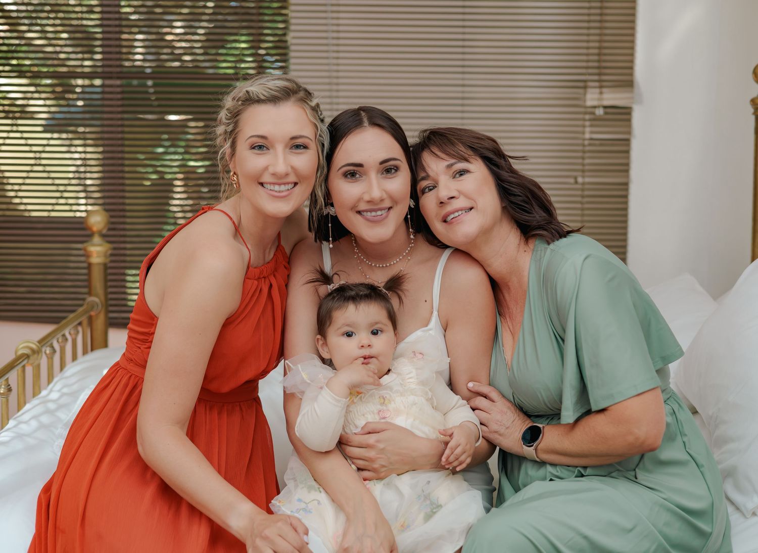 Bride holding infant smiling with bridesmaid in orange dress and older woman in sage green outfit.