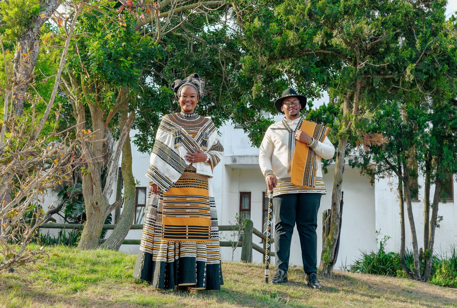 Two people in traditional Xhosa attire stand outdoors holding musical instruments near trees and a white building.