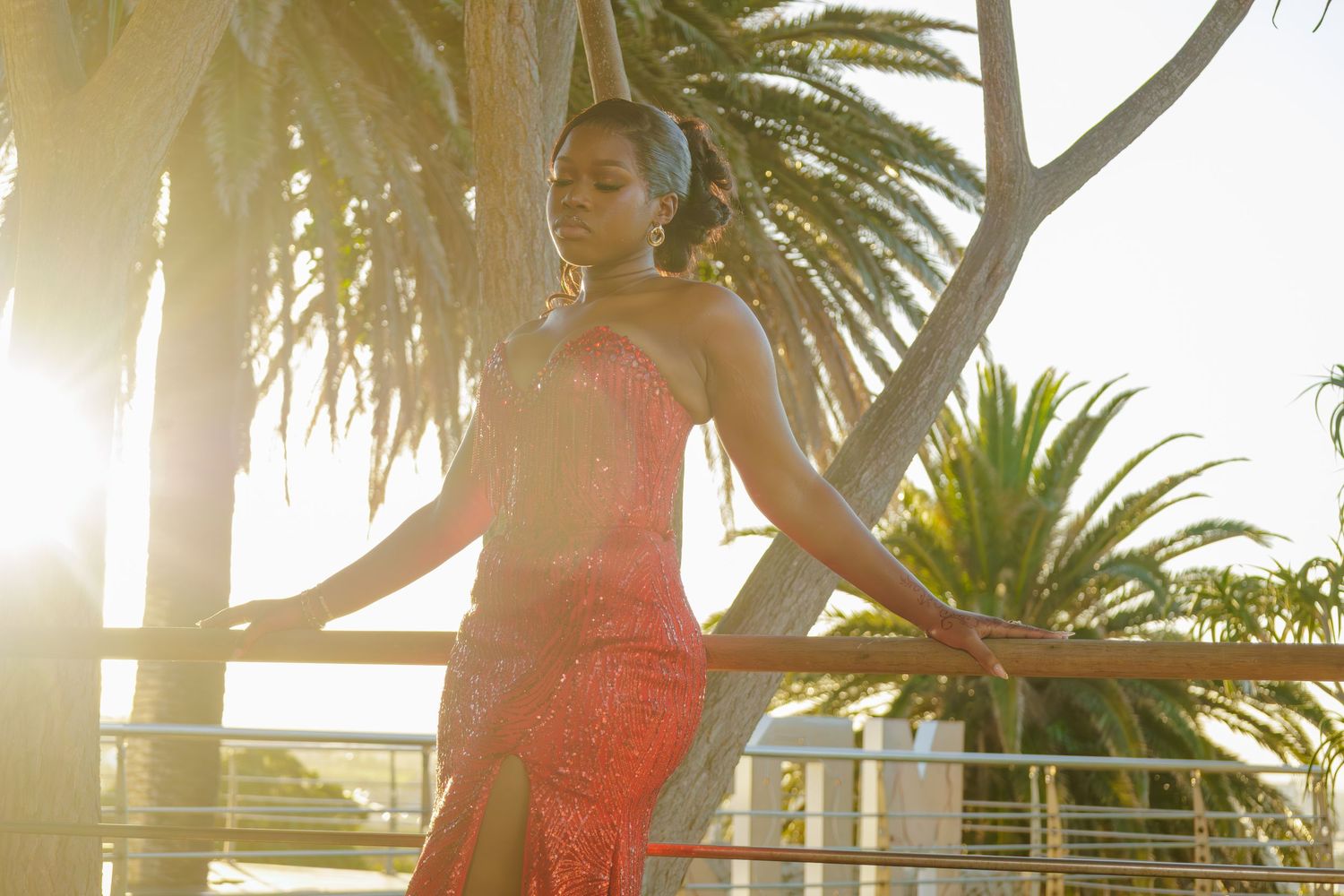 A woman in a red lace gown poses gracefully by a railing with palm trees and golden sunlight behind her.