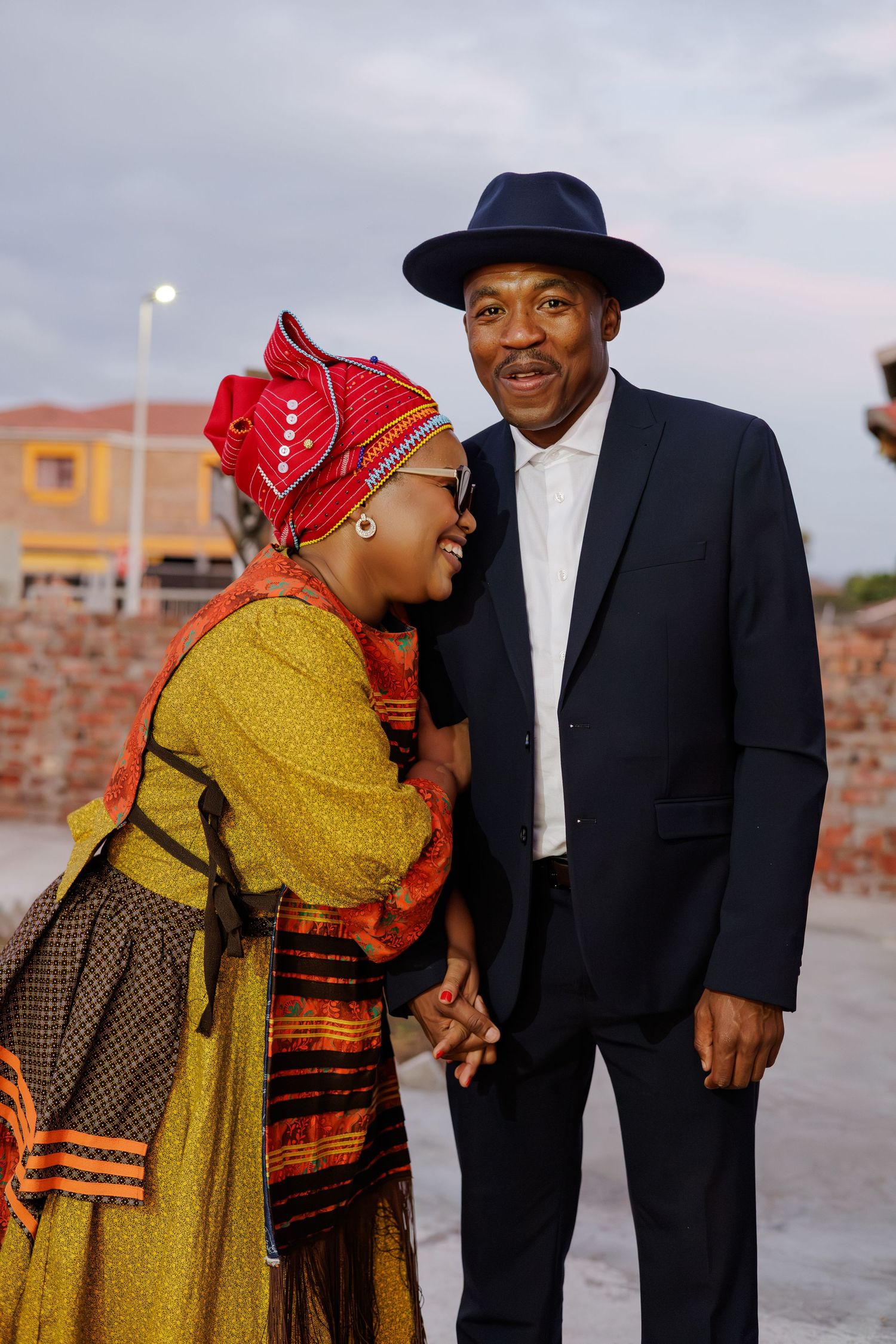 Couple in traditional African attire holding hands outdoors, woman in colorful dress and headwrap, man in navy suit and hat.