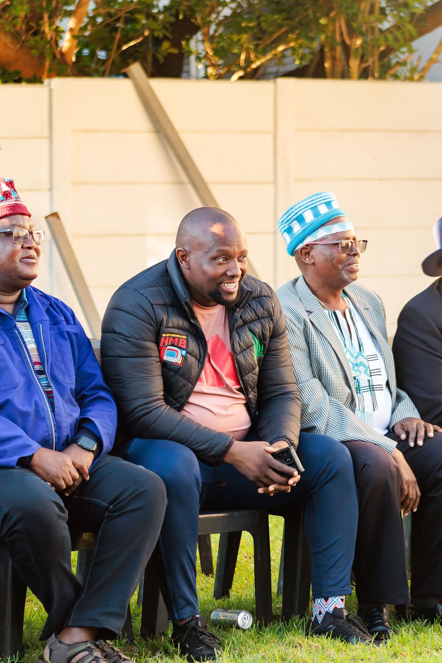 Three men in traditional African caps sit smiling outdoors at a cultural gathering.