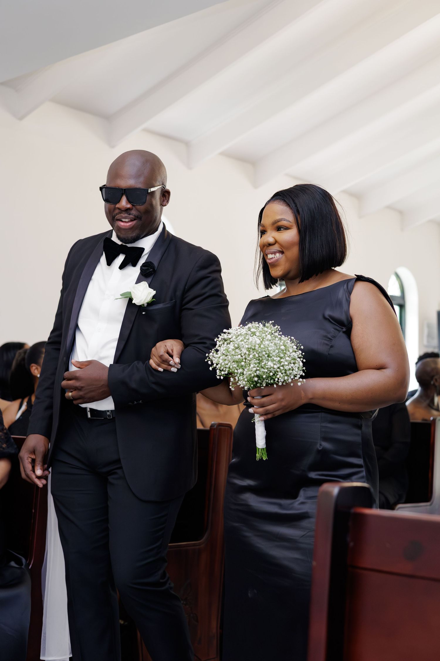 Smiling groomsman in sunglasses escorts bridesmaid in black dress down church aisle.