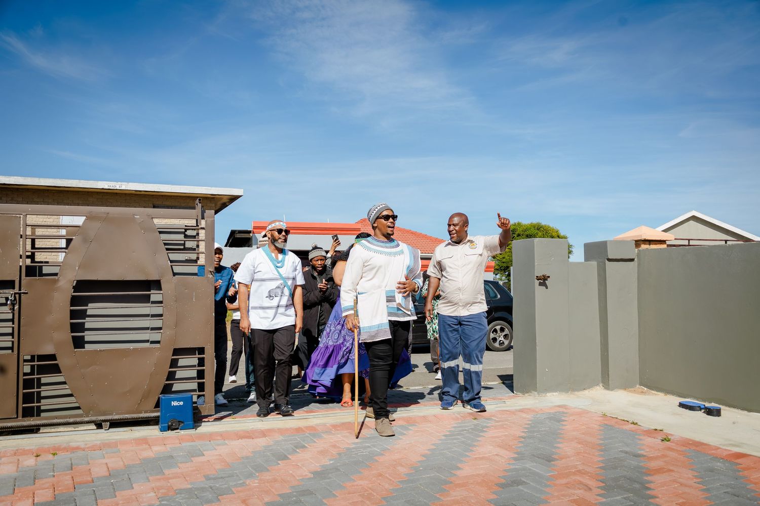 A group of people walk through open gates onto a brick pathway under a bright blue sky.