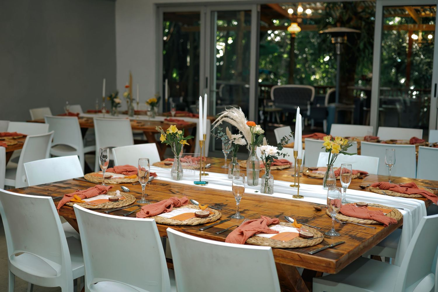 Elegantly set rustic wooden tables with terracotta napkins, gold chargers, and floral centerpieces in a bright wedding reception venue.