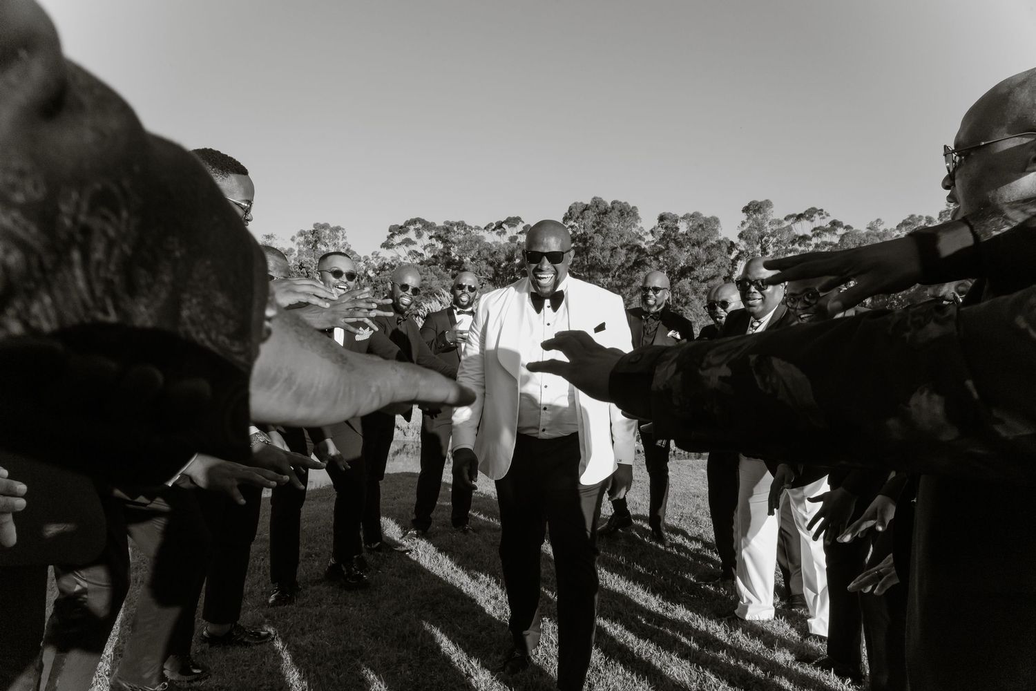 Groom in white tuxedo greeted by cheering wedding guests in a dramatic black and white outdoor photo.