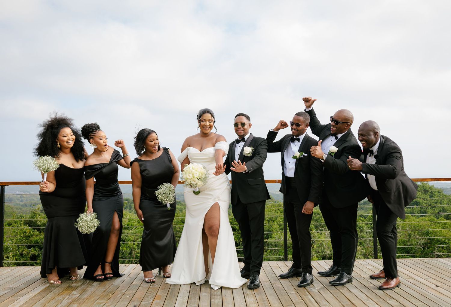 Joyful wedding party in black attire celebrating with bride and groom on a scenic outdoor deck.