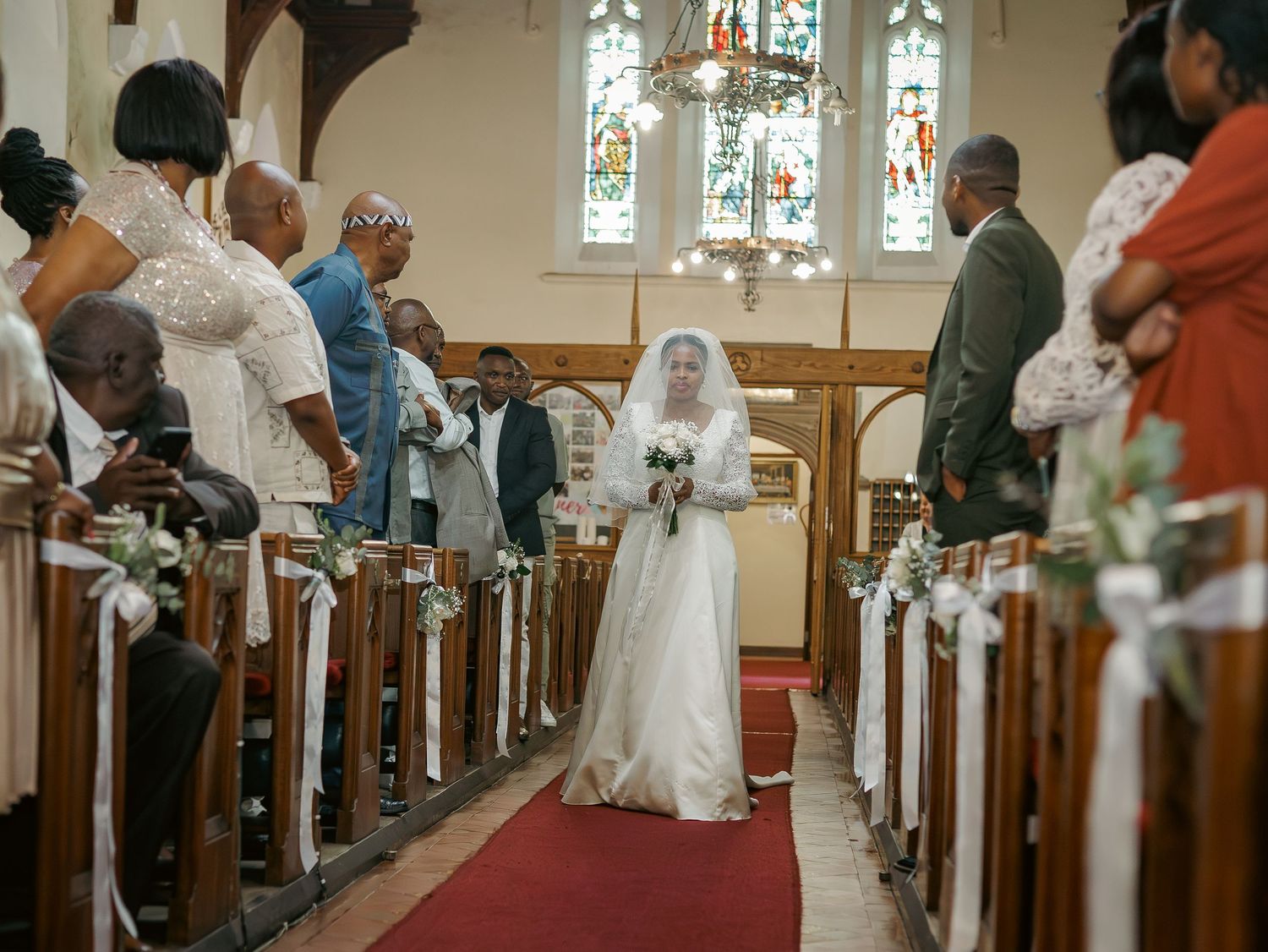 Bride walks down red carpet aisle in white gown past seated wedding guests.