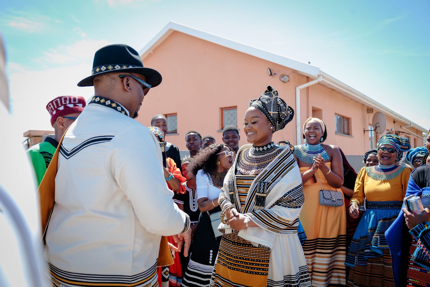 Groom in white Xhosa attire and black hat holds hands with bride in traditional beaded dress outdoors.