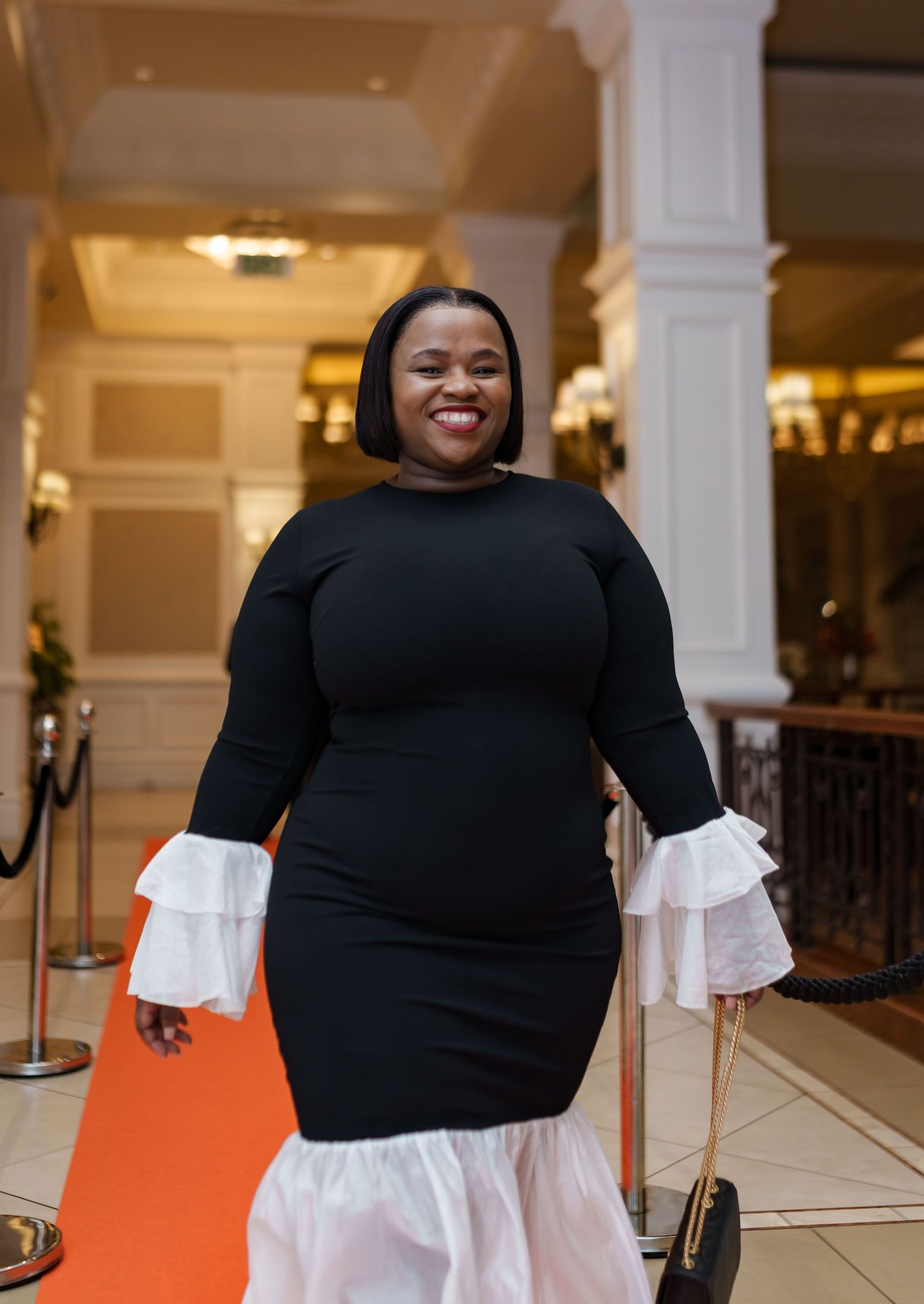 Woman in black ruffle dress smiling on orange carpet at elegant corporate event.
