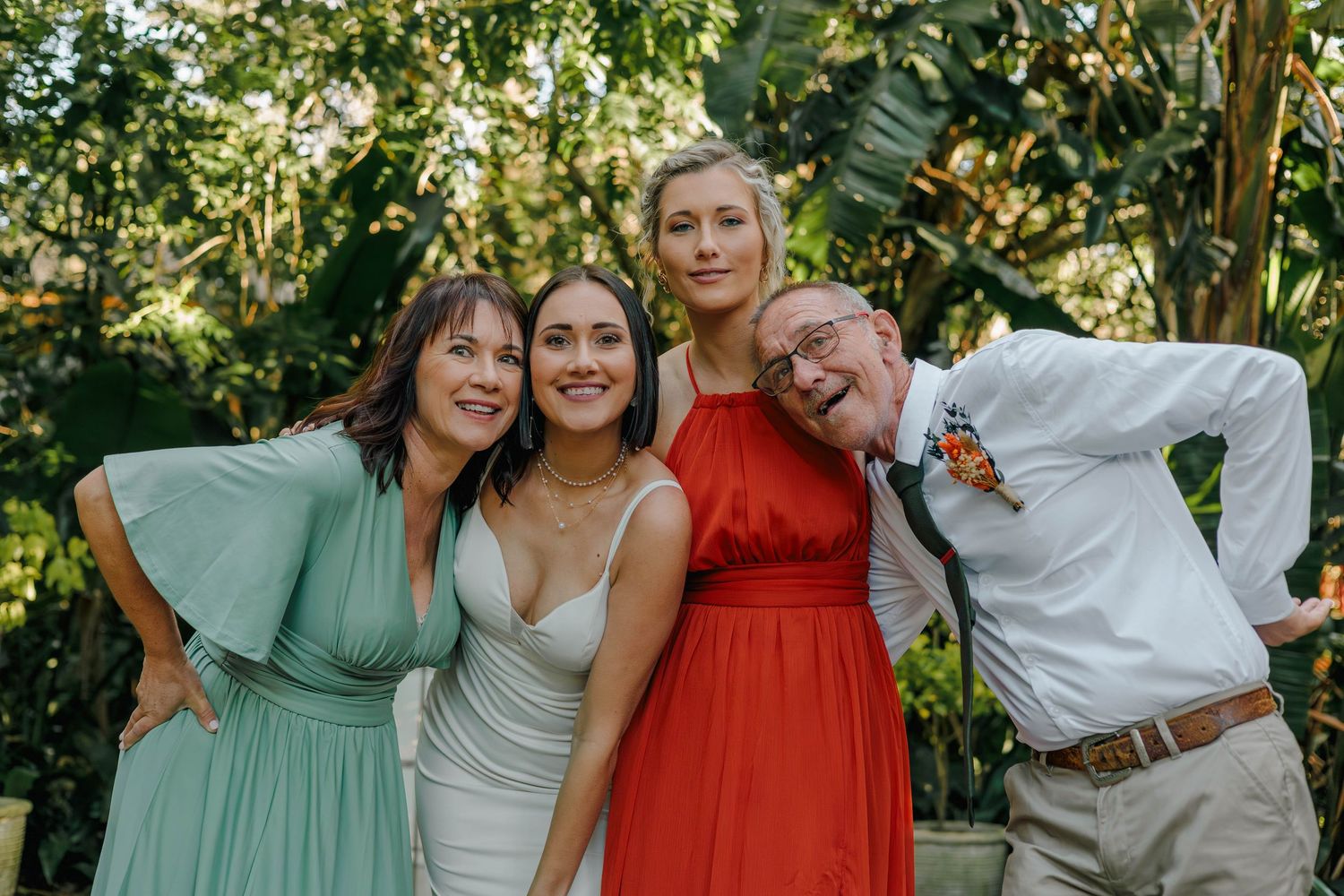 Bride smiles with family members in mint, red dresses and suit in tropical garden.