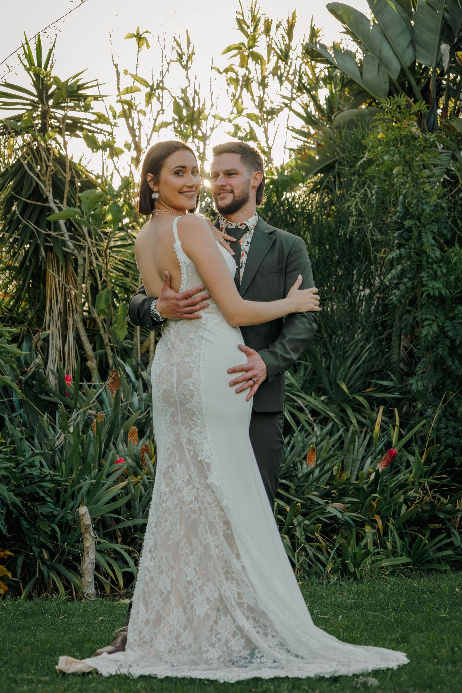 Bride in lace gown and groom embrace in front of lush tropical garden at sunset.